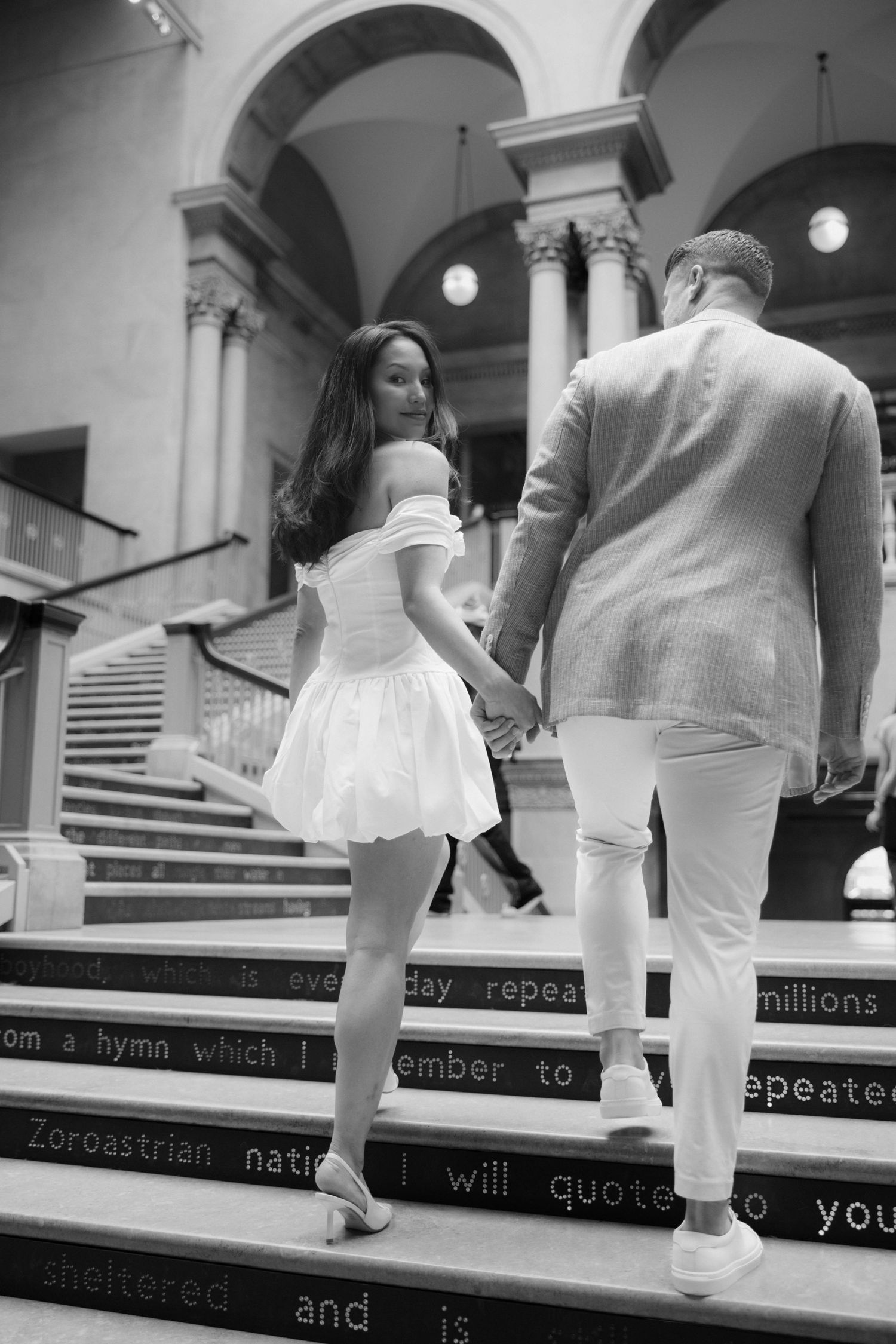 Black and white photo of a couple holding hands walking up grand staircase, woman in white dress, man in light suit.