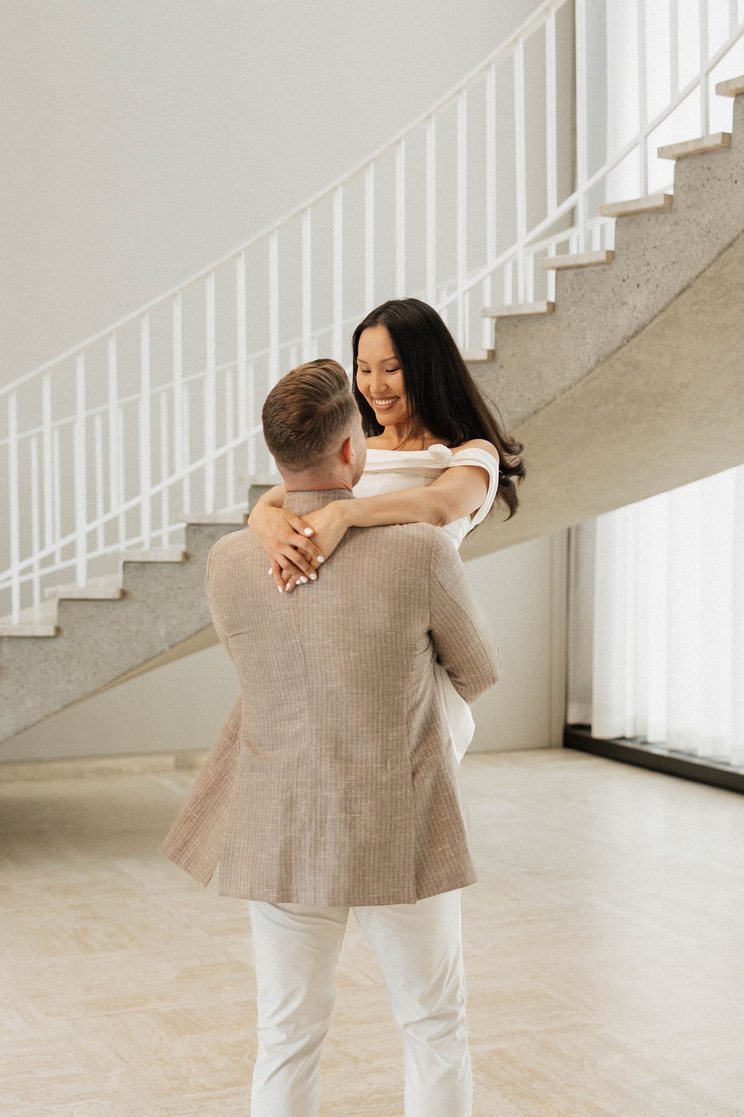 A couple dances together in an elegant white room with a spiral staircase, the woman smiling warmly in a white dress.