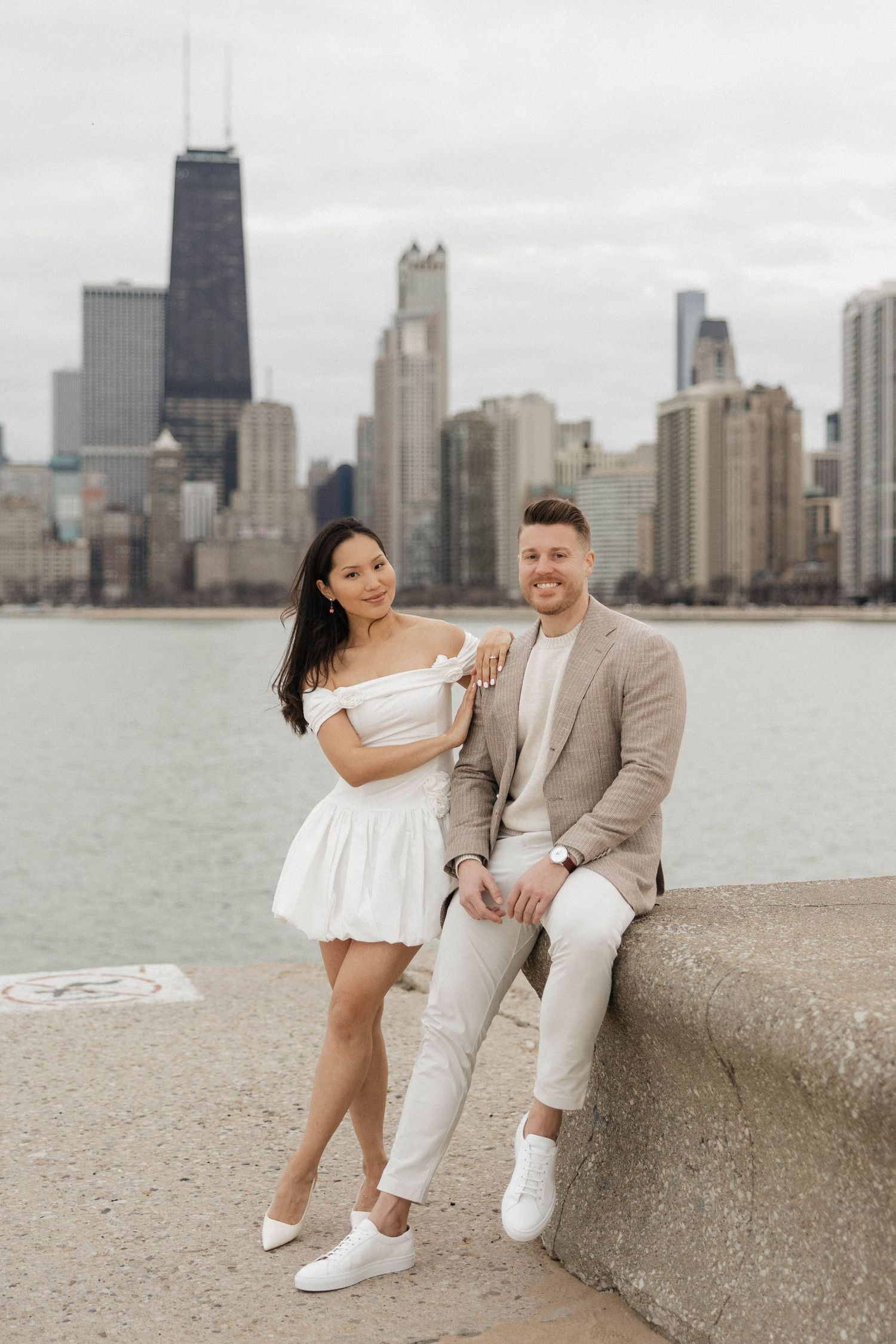 Couple poses by Lake Michigan with Chicago skyline backdrop, woman in white dress, man in beige suit, smiling.