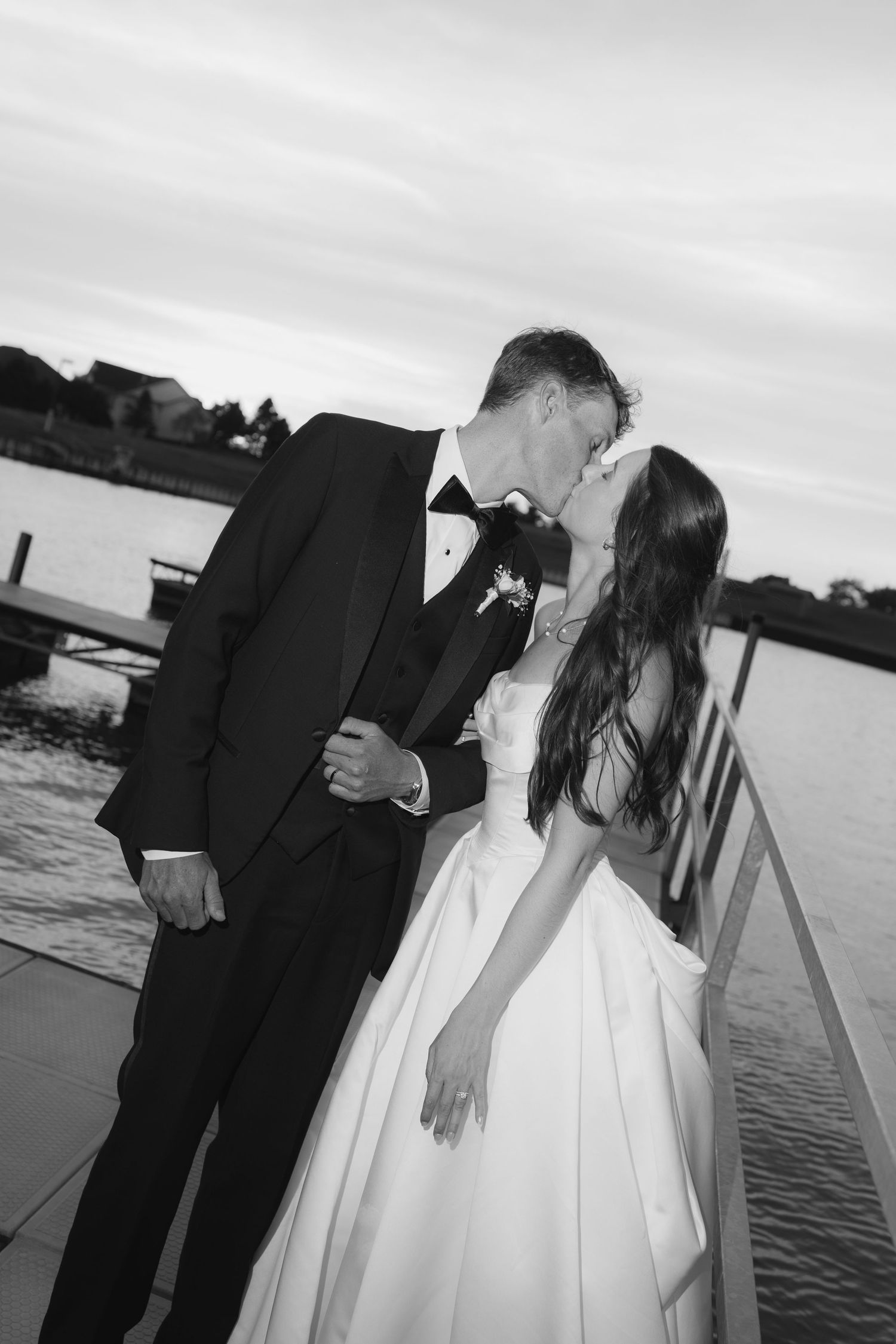 Black and white photo of a bride and groom sharing a kiss on a waterfront dock at sunset.