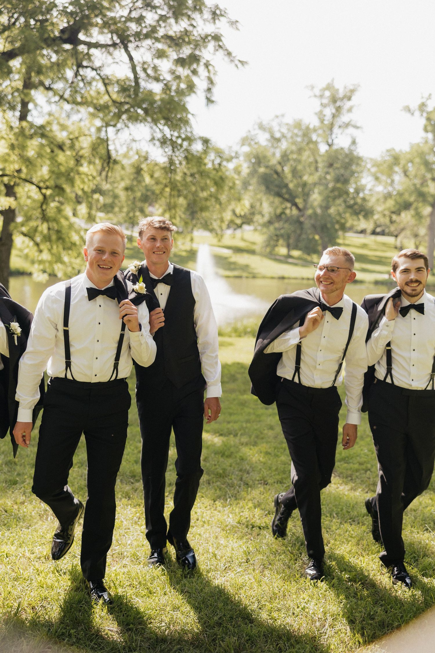 Groomsmen in white shirts, suspenders, and bow ties walk outdoors near a pond, some carrying jackets, on a sunny wedding day.