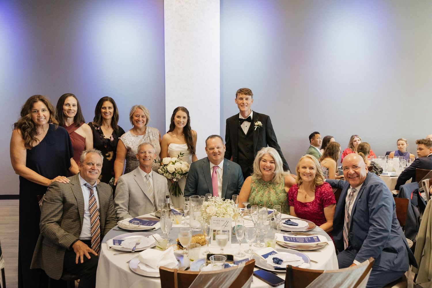 Group of elegantly dressed family members posing together at a wedding reception dinner table with floral centerpiece.