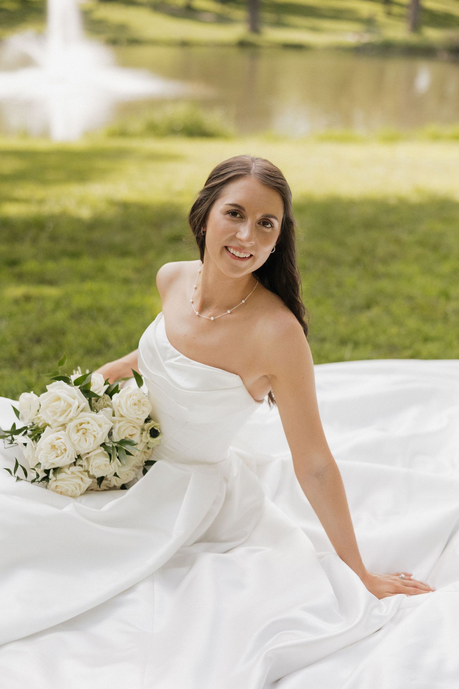 Smiling bride in white strapless gown sitting on grass holding white rose bouquet near a pond.