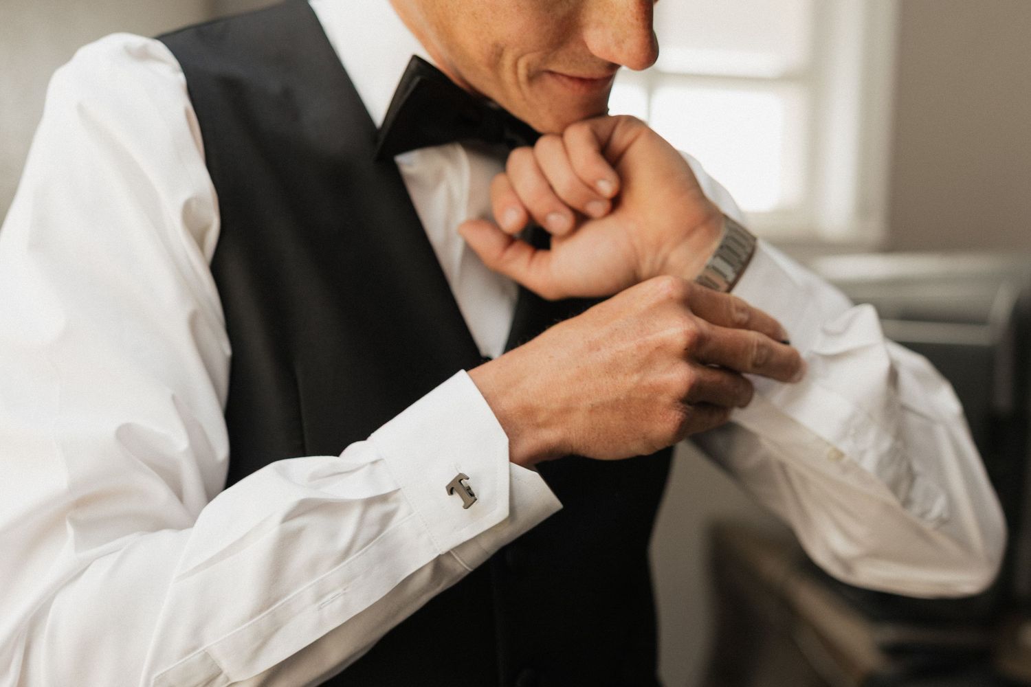 Man in formal black vest and bow tie fastening silver cufflinks on white dress shirt sleeves.