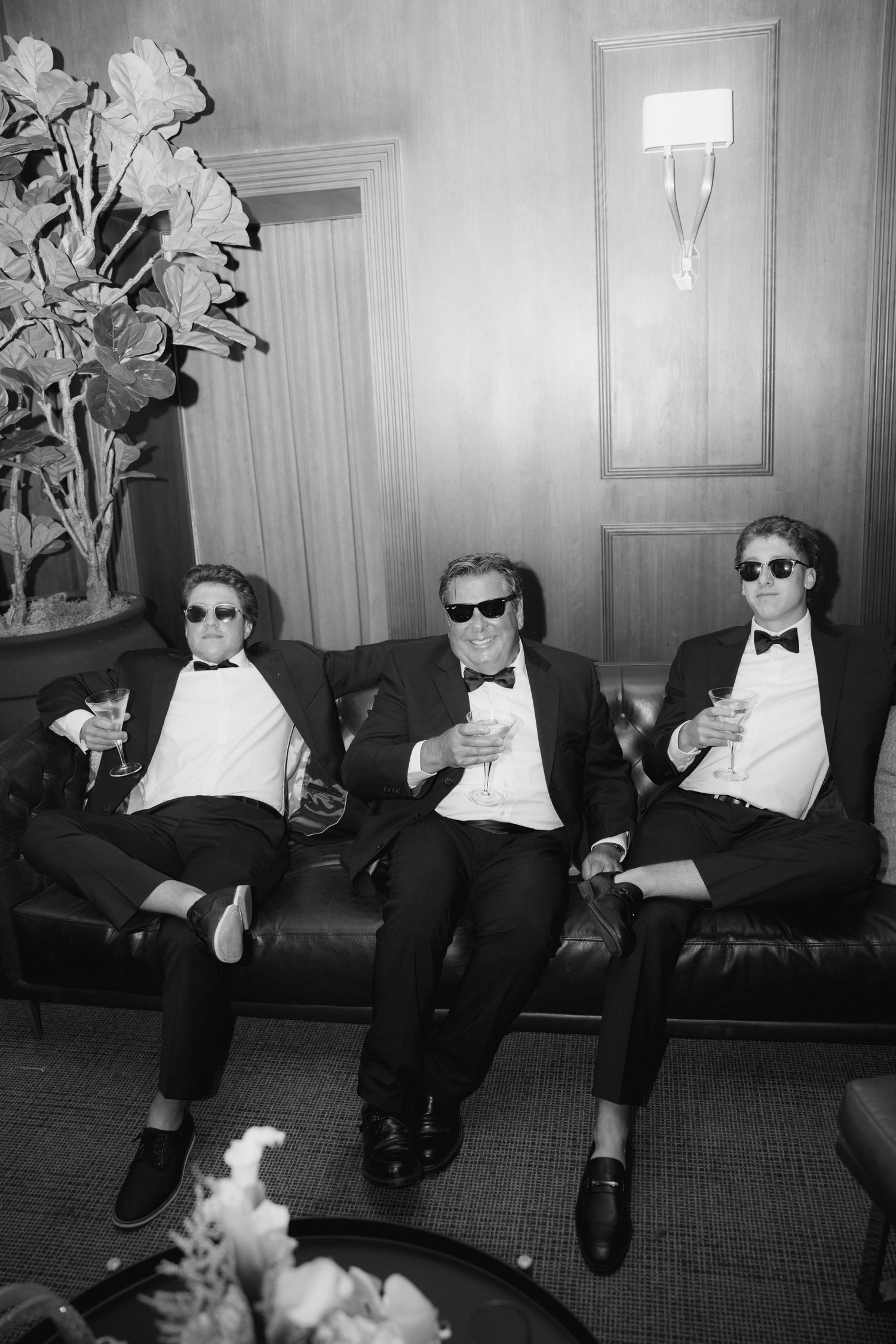 Three men in tuxedos and sunglasses sit on a leather sofa holding champagne glasses in a black and white photo.