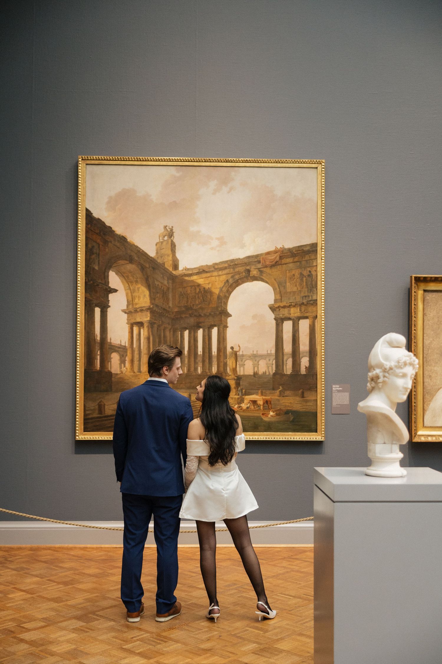 Couple viewing classical paintings in an art museum gallery with wooden floors.