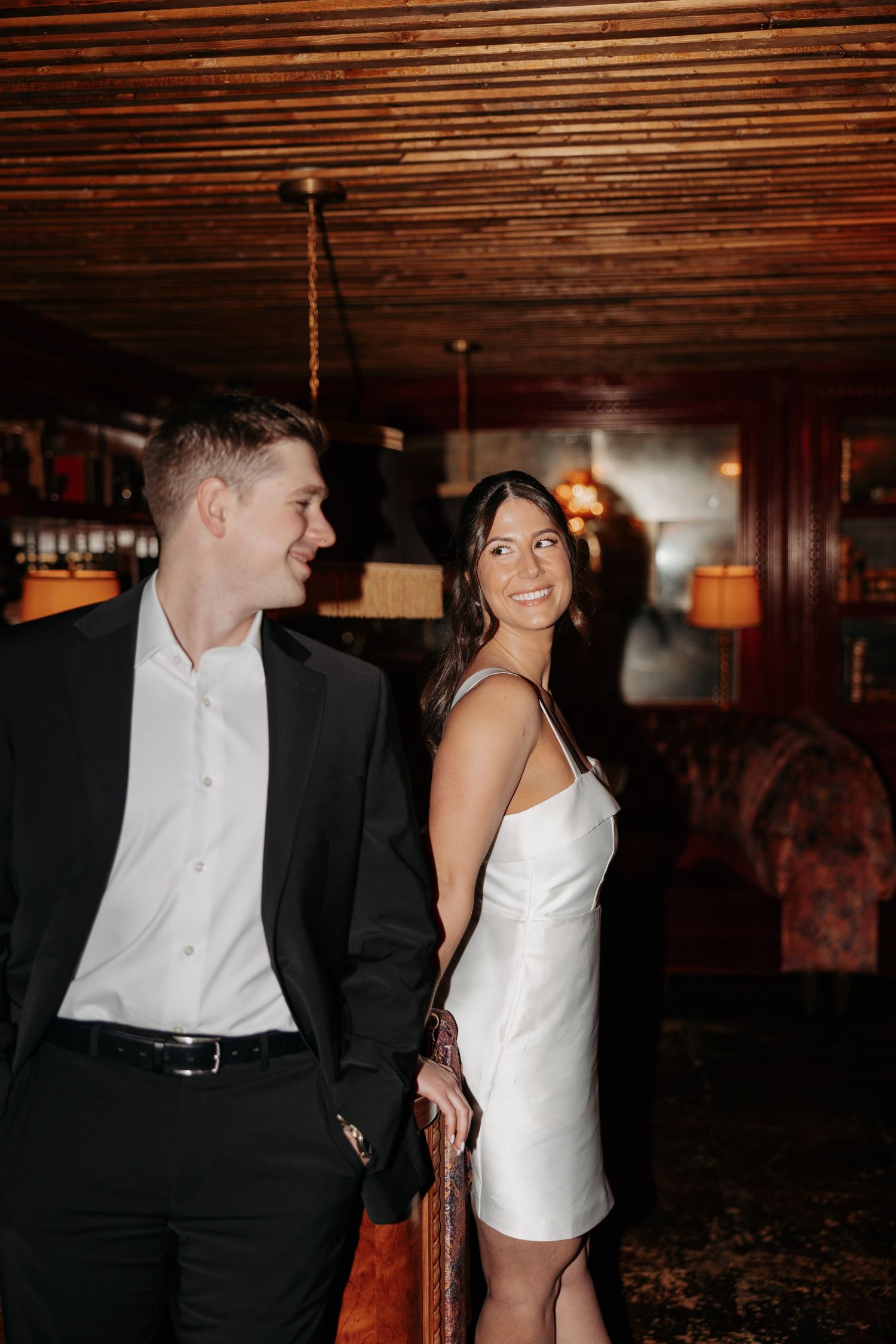 A couple in formal attire share a moment in a dimly lit room with wooden walls and warm lighting.
