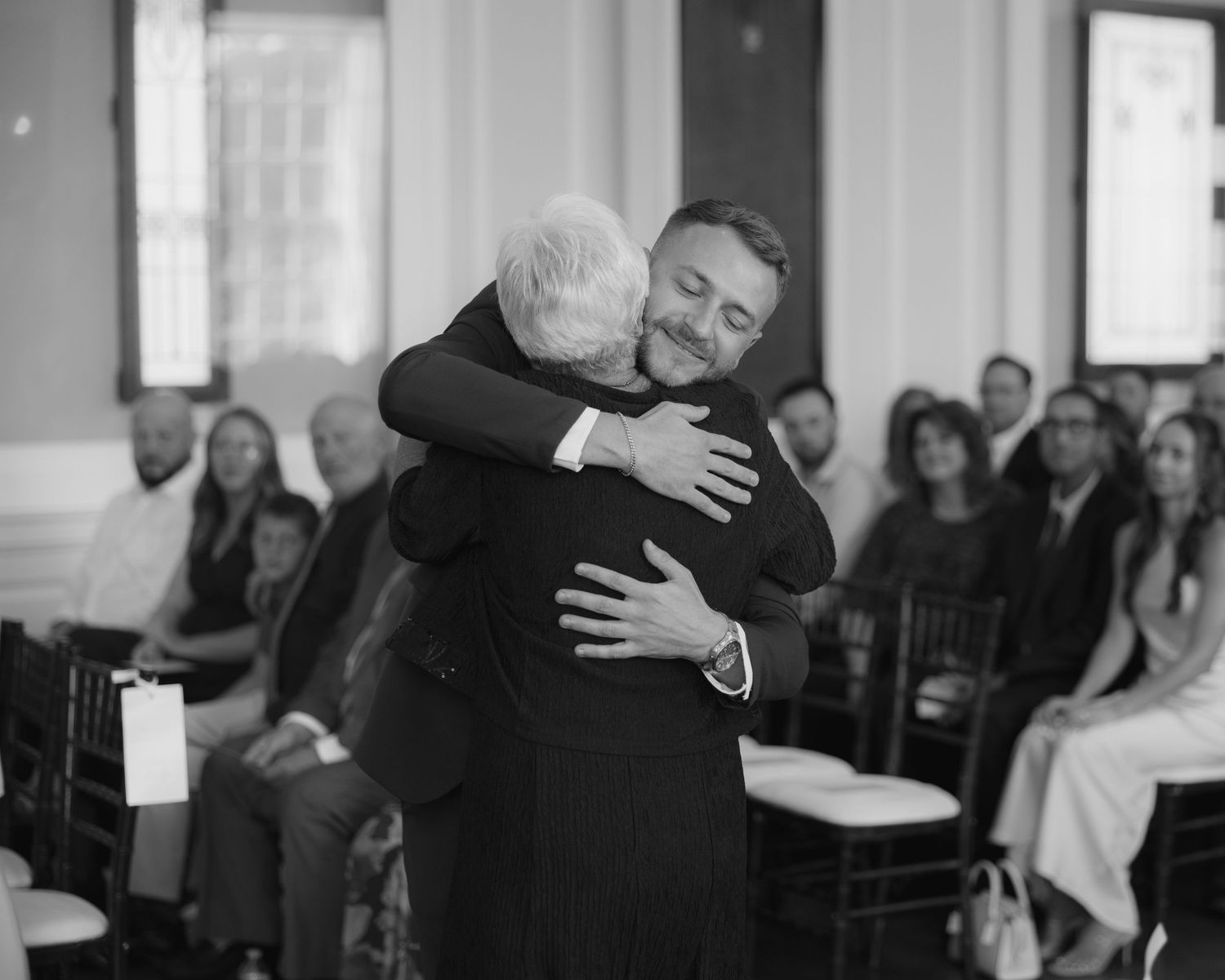 Two people embrace emotionally during a wedding ceremony as onlookers watch from their seats in a bright indoor venue.