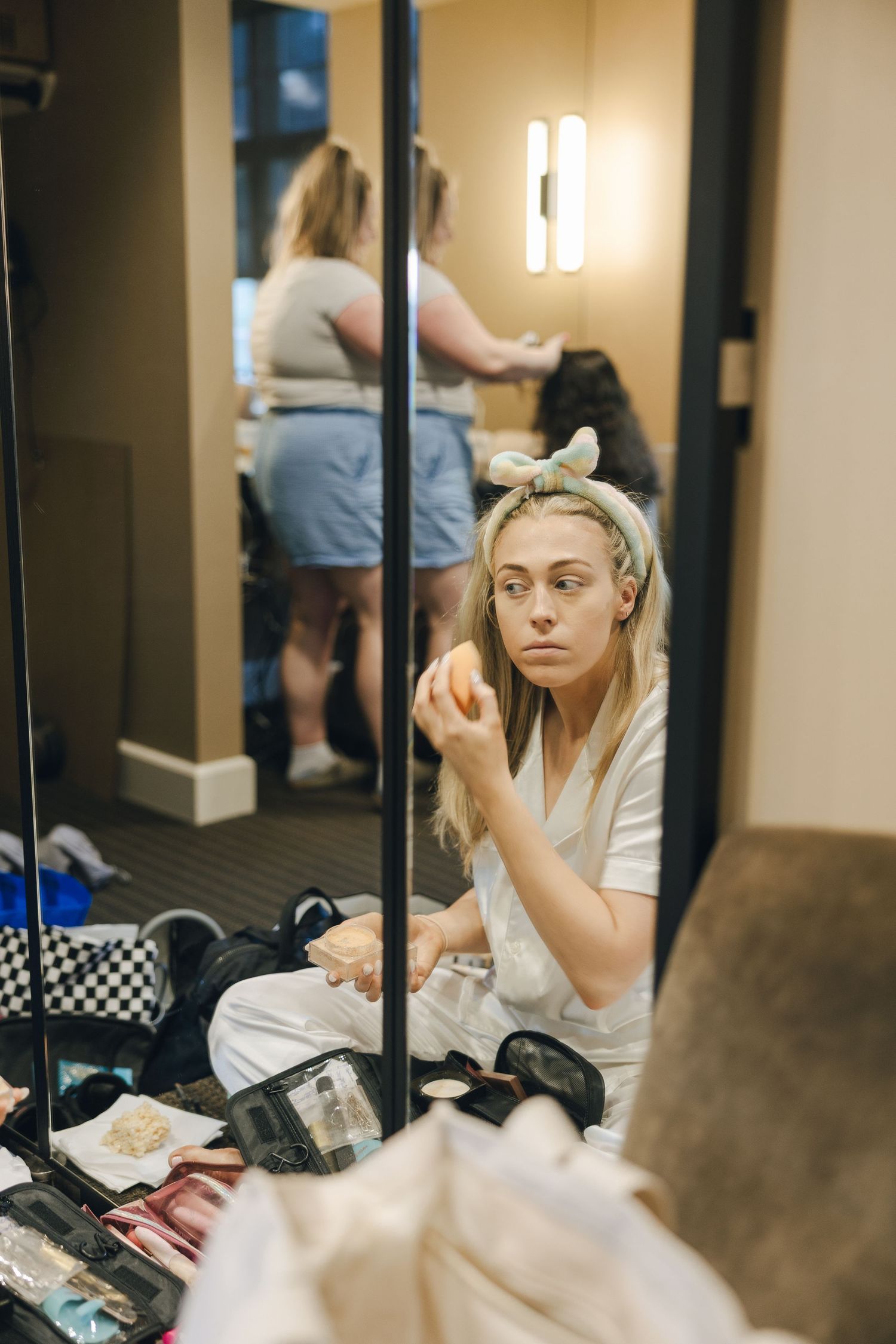 Backstage dressing room scene with makeup and personal items scattered on counter while someone prepares in mirror.