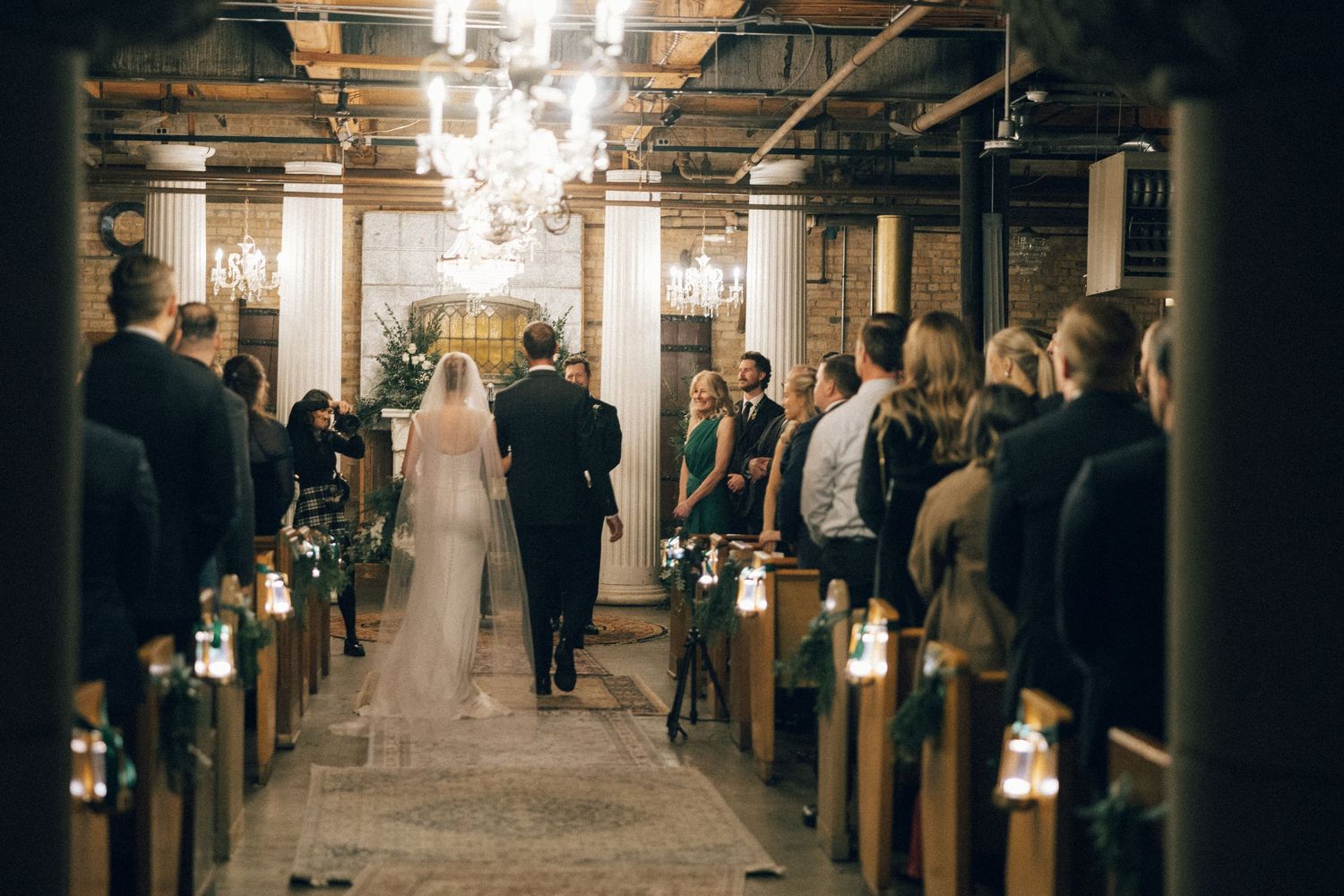 Wedding ceremony in a rustic venue with wooden pews and glowing lanterns lining the aisle.