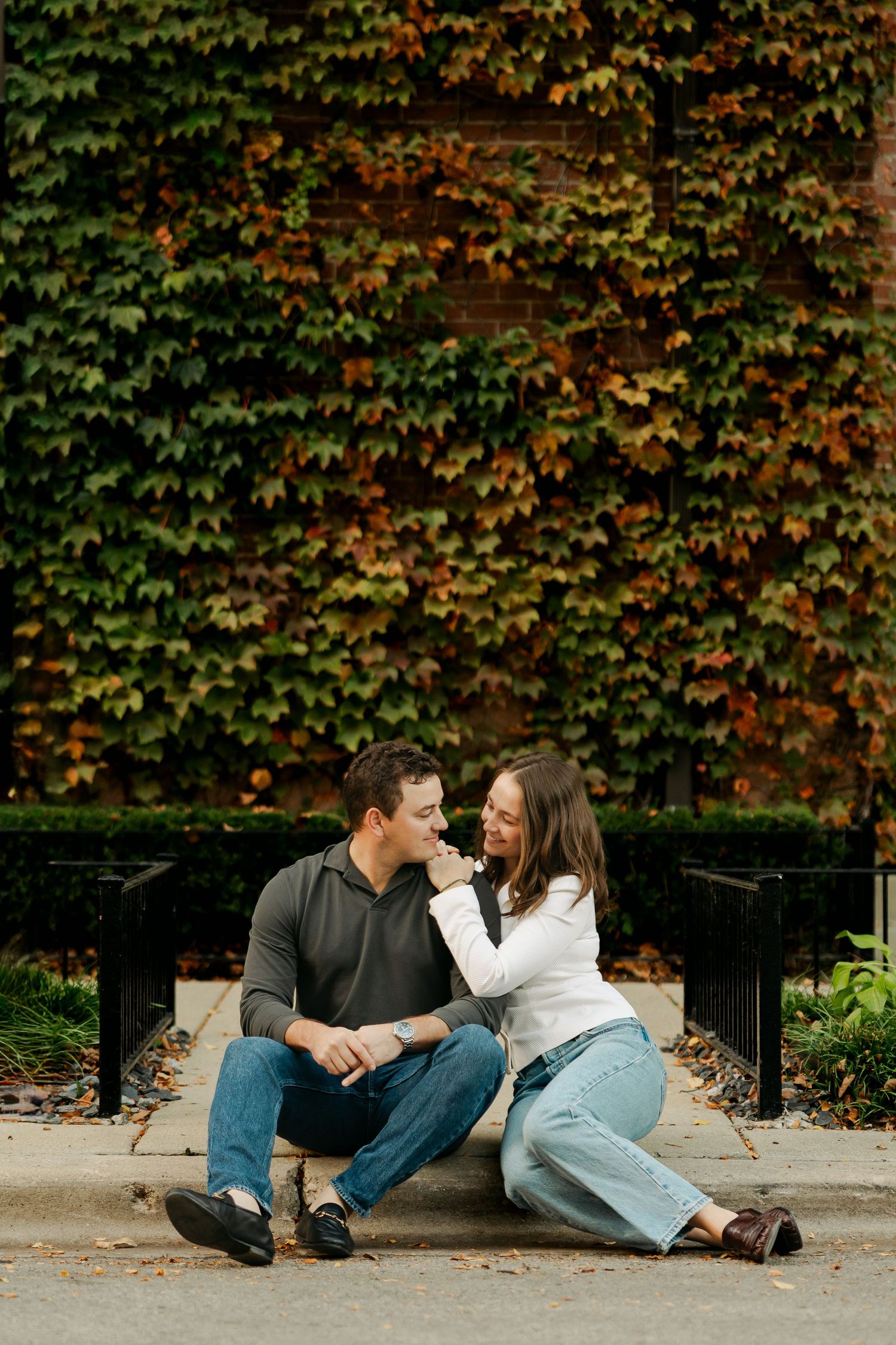 Smiling couple sitting close on a city curb in front of a fall ivy-covered brick wall, perfect for an engagement photo session.