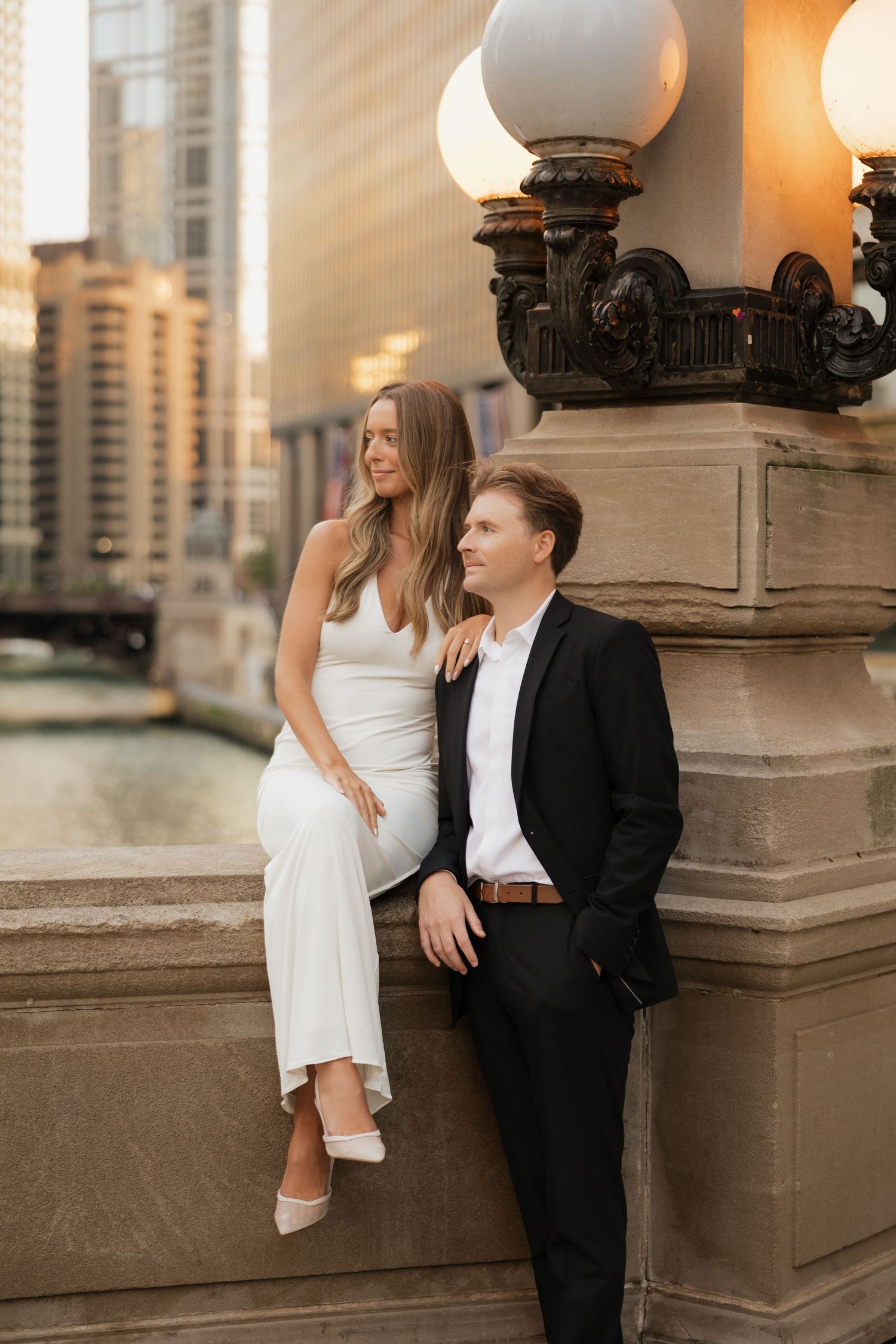 Couple in white dress and black suit pose on a stone ledge beside an ornate lamp post with a city river and skyscrapers behind.