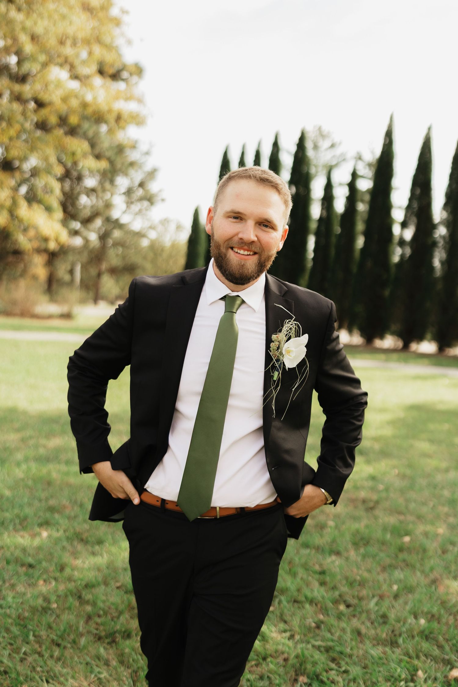 Smiling groom in a black suit, green tie and floral boutonniere stands with hands in pockets on a grassy lawn lined with cypress trees.