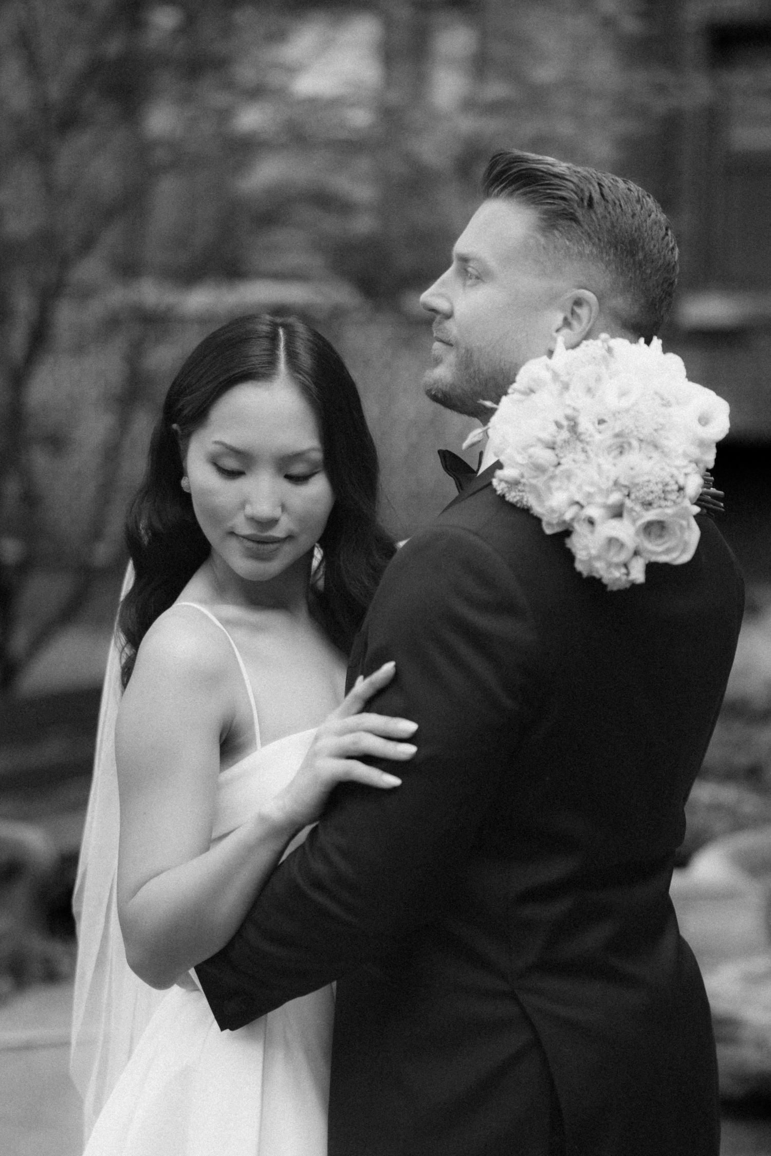 Black and white wedding portrait of a bride in a spaghetti-strap gown and veil embracing a groom in a tuxedo holding a white rose bouquet.