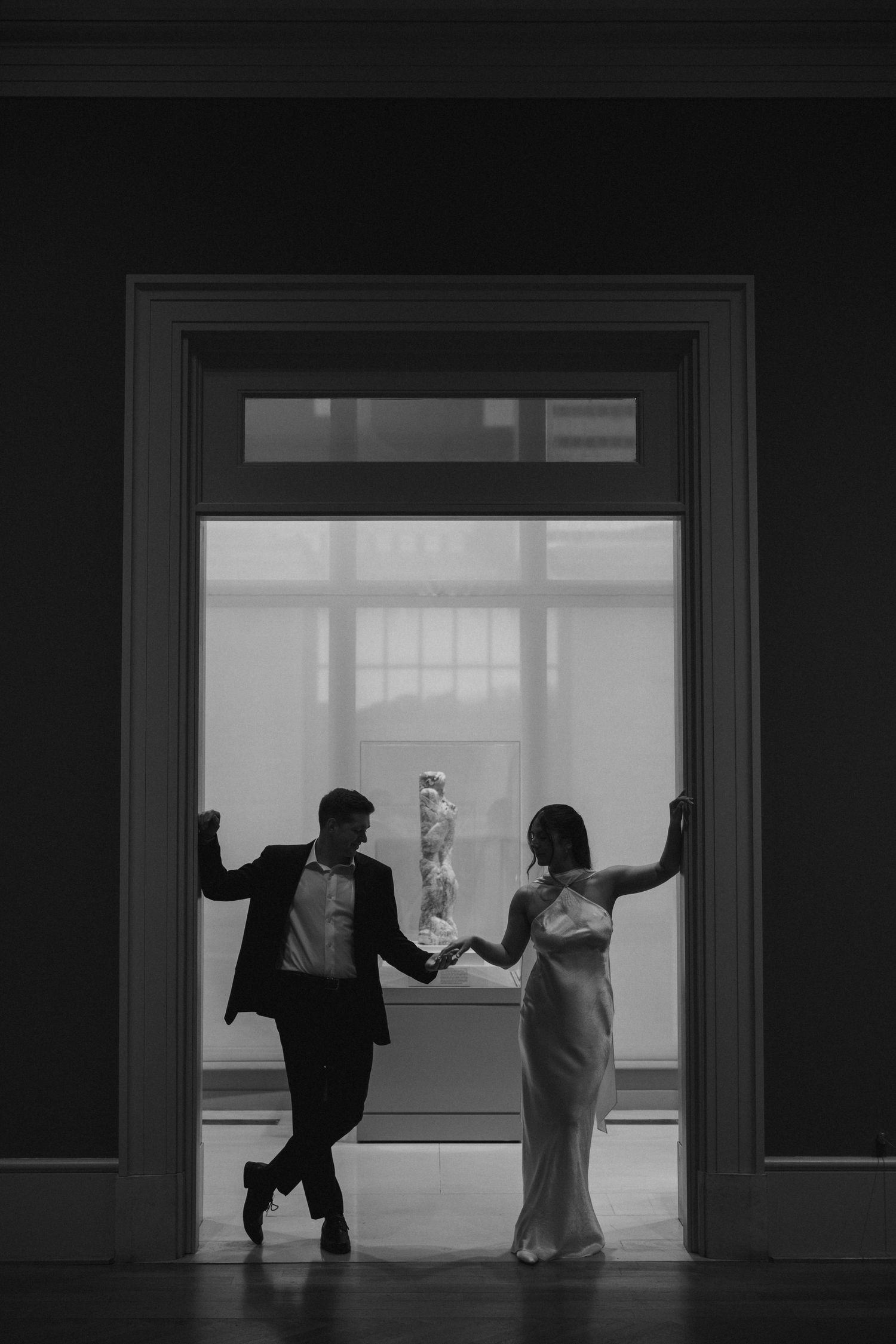 Black and white photo of a couple holding hands in a doorway, with a classical statue visible behind them.
