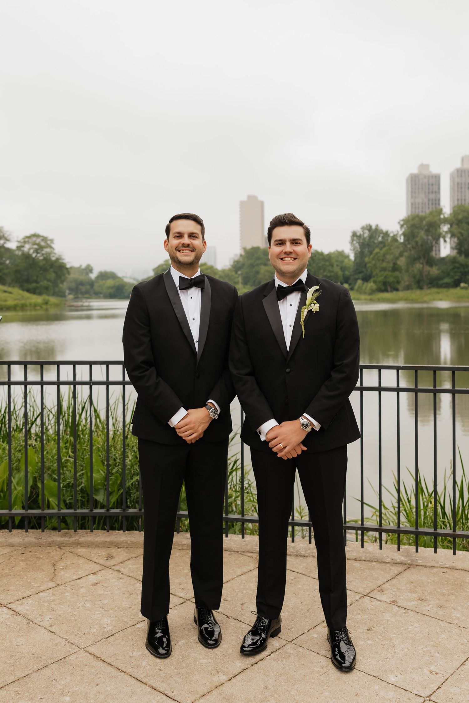 Two grooms in black tuxedos smile outdoors by a lake with city skyline, surrounded by lush greenery.