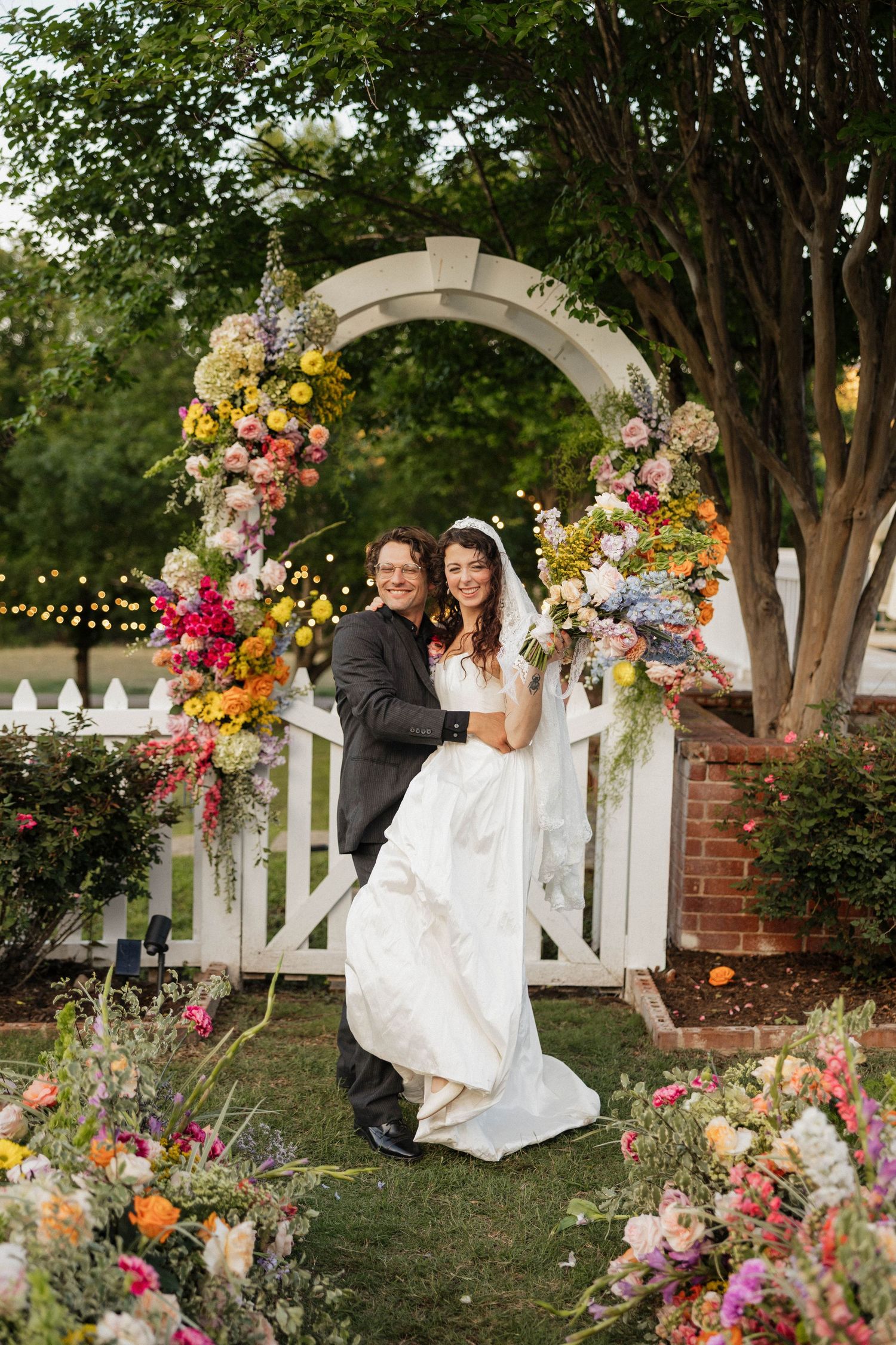 Bride and groom pose under a colorful floral arch at an outdoor wedding ceremony surrounded by vibrant blooms.
