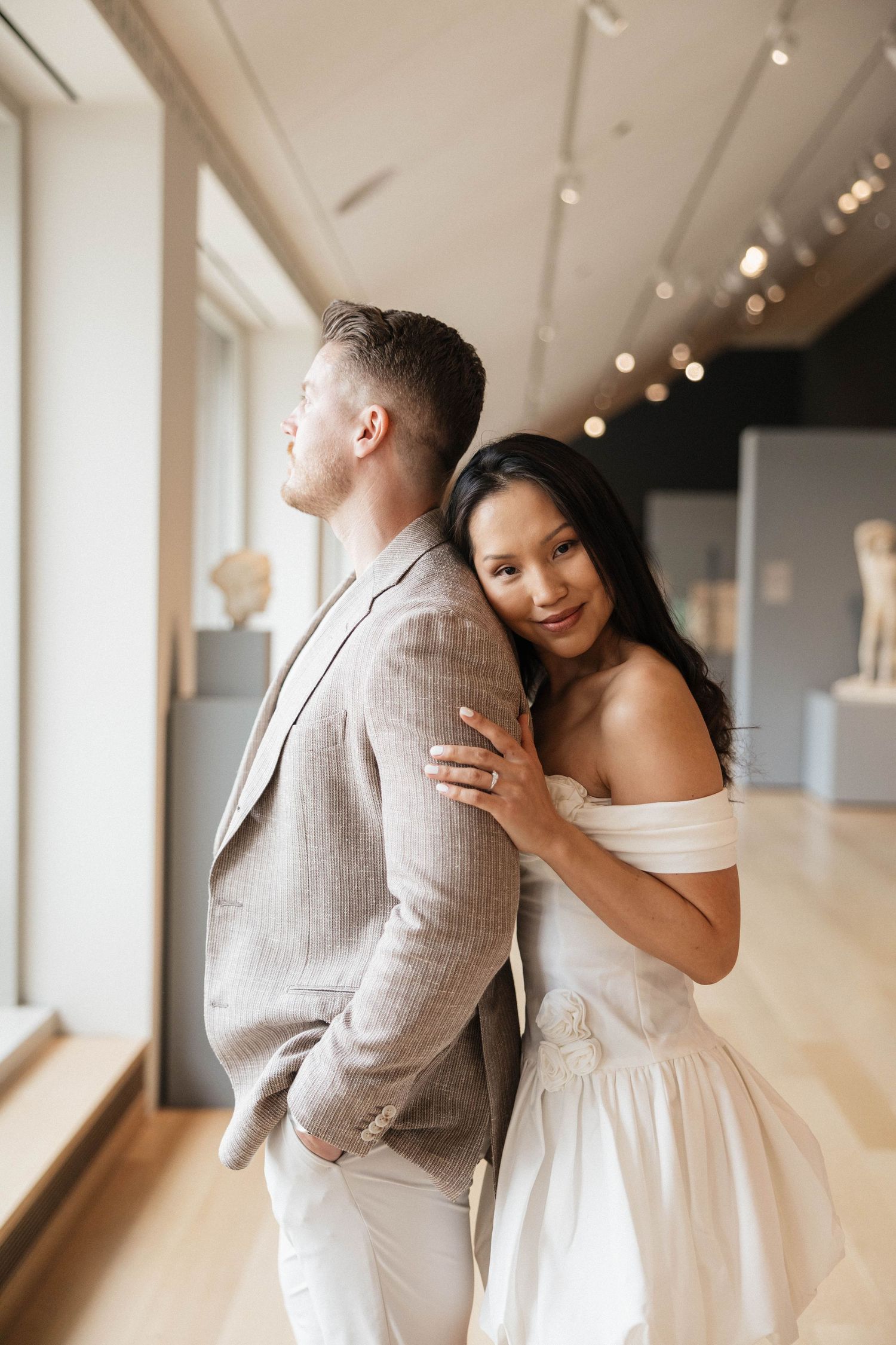 Elegant couple posing in art museum, bride in off-shoulder white dress leaning on groom in linen suit.