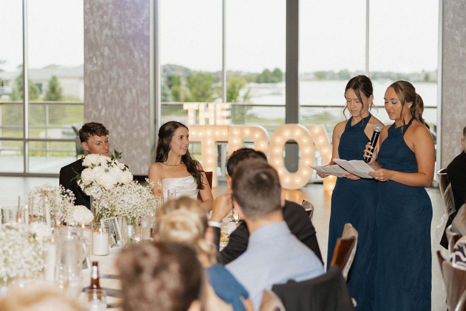 Two bridesmaids in navy dresses give a wedding speech at an elegant reception venue with large windows.