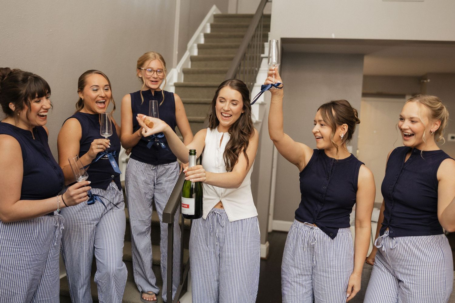 Six bridesmaids in navy tops and striped pants celebrate with champagne, laughing joyfully before a wedding ceremony.