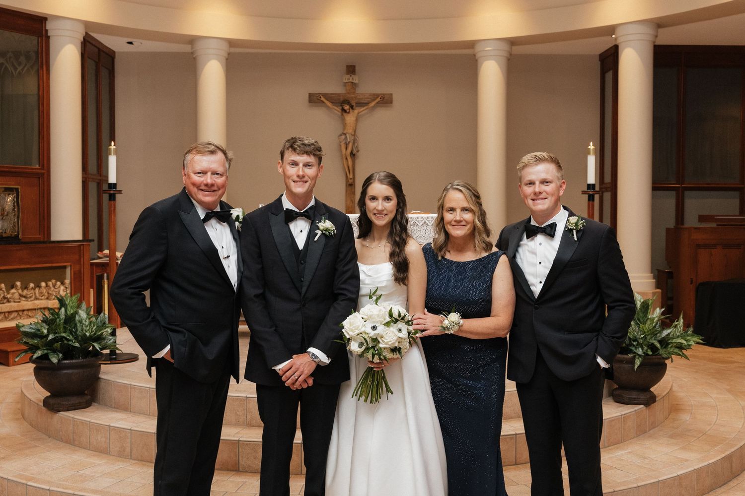 Wedding party poses before altar with crucifix in elegant church, bride holding white bouquet flanked by family in formal attire.