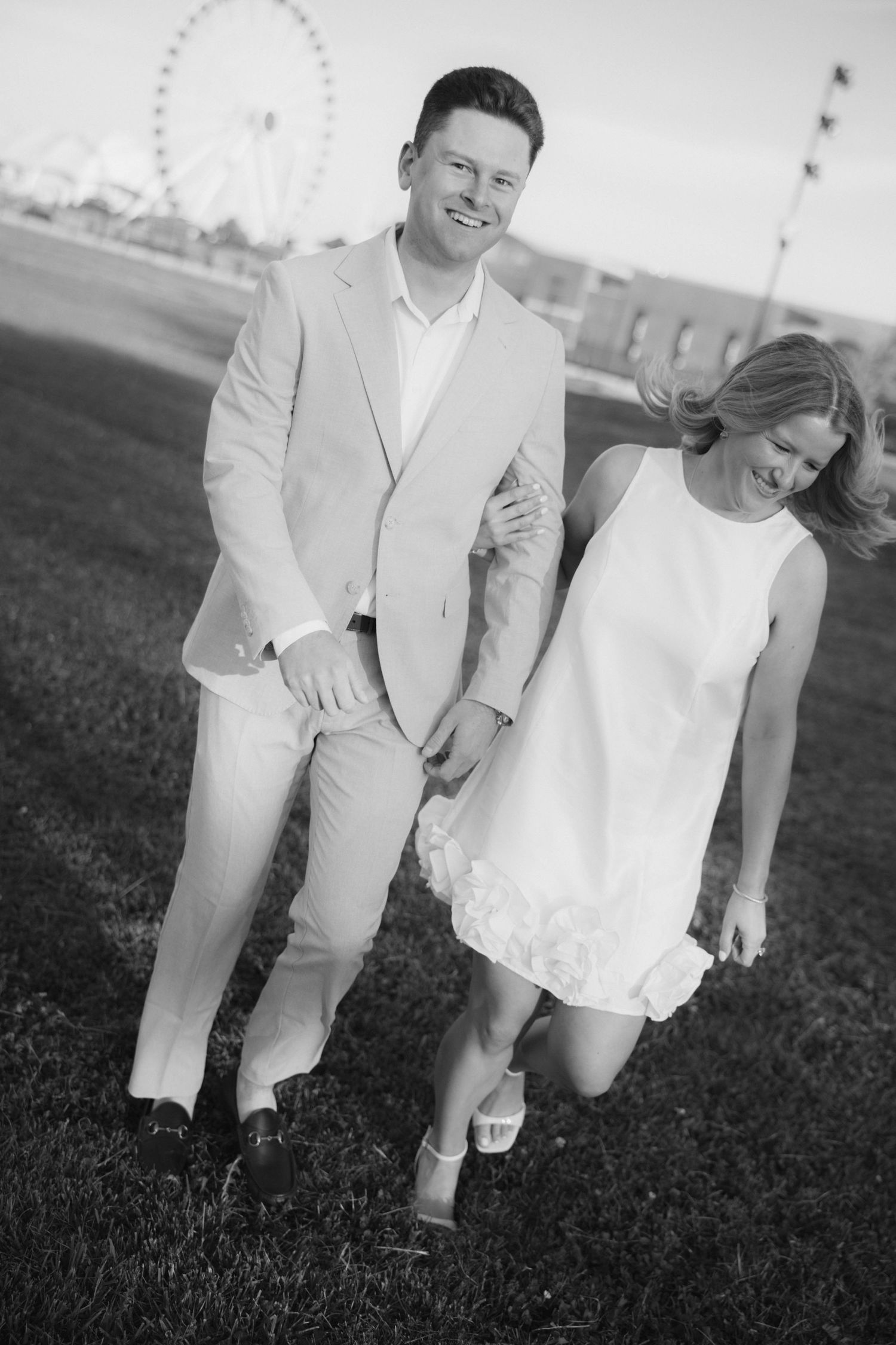 Happy couple walking on grass with Ferris wheel backdrop in black and white engagement photo.