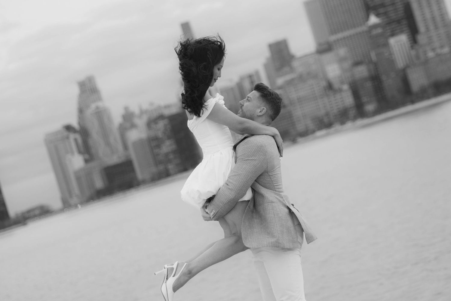 Black and white photo of a man lifting a woman in a white dress with a city skyline behind them.