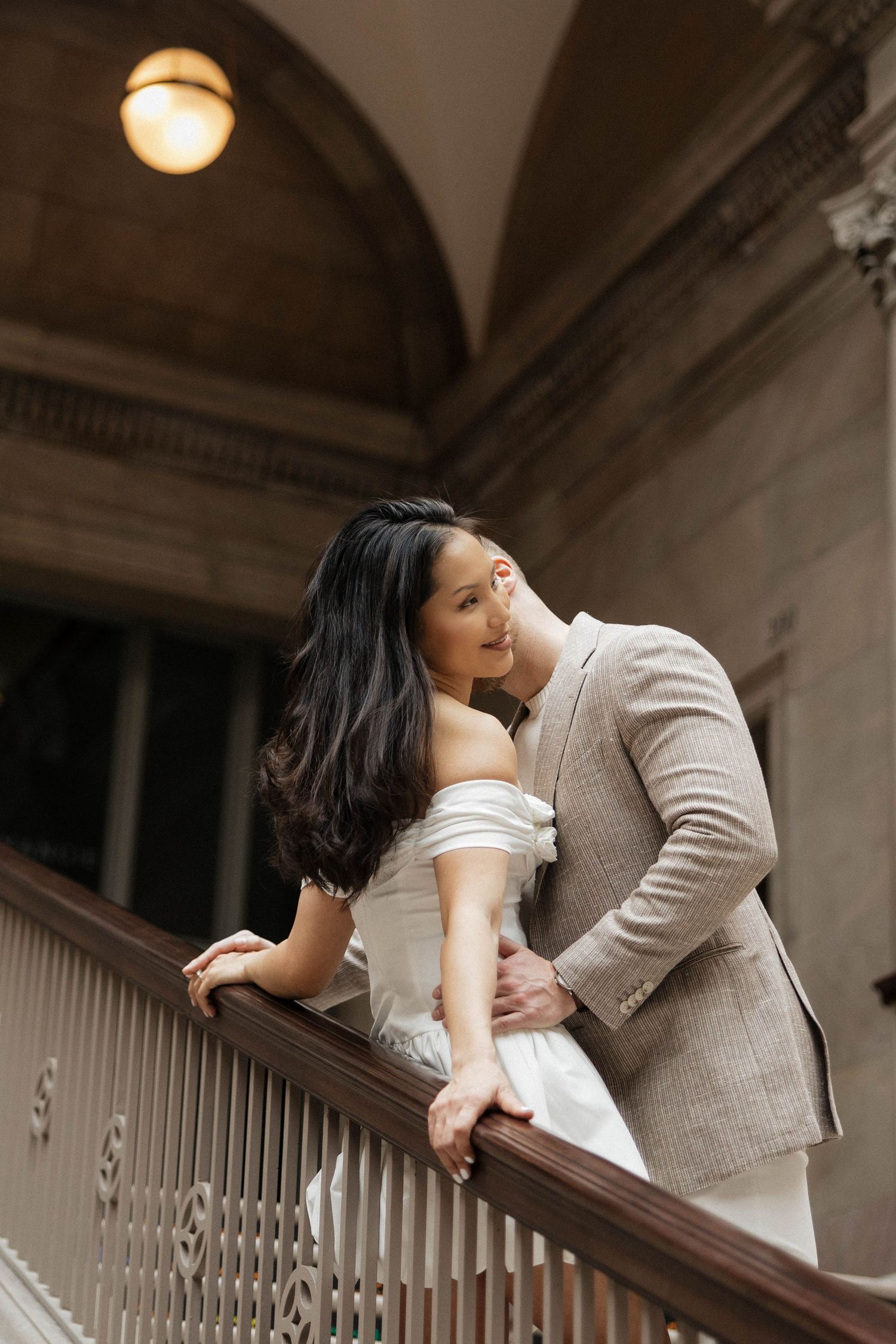 Couple in elegant wedding attire embracing on ornate staircase in classic architectural interior.