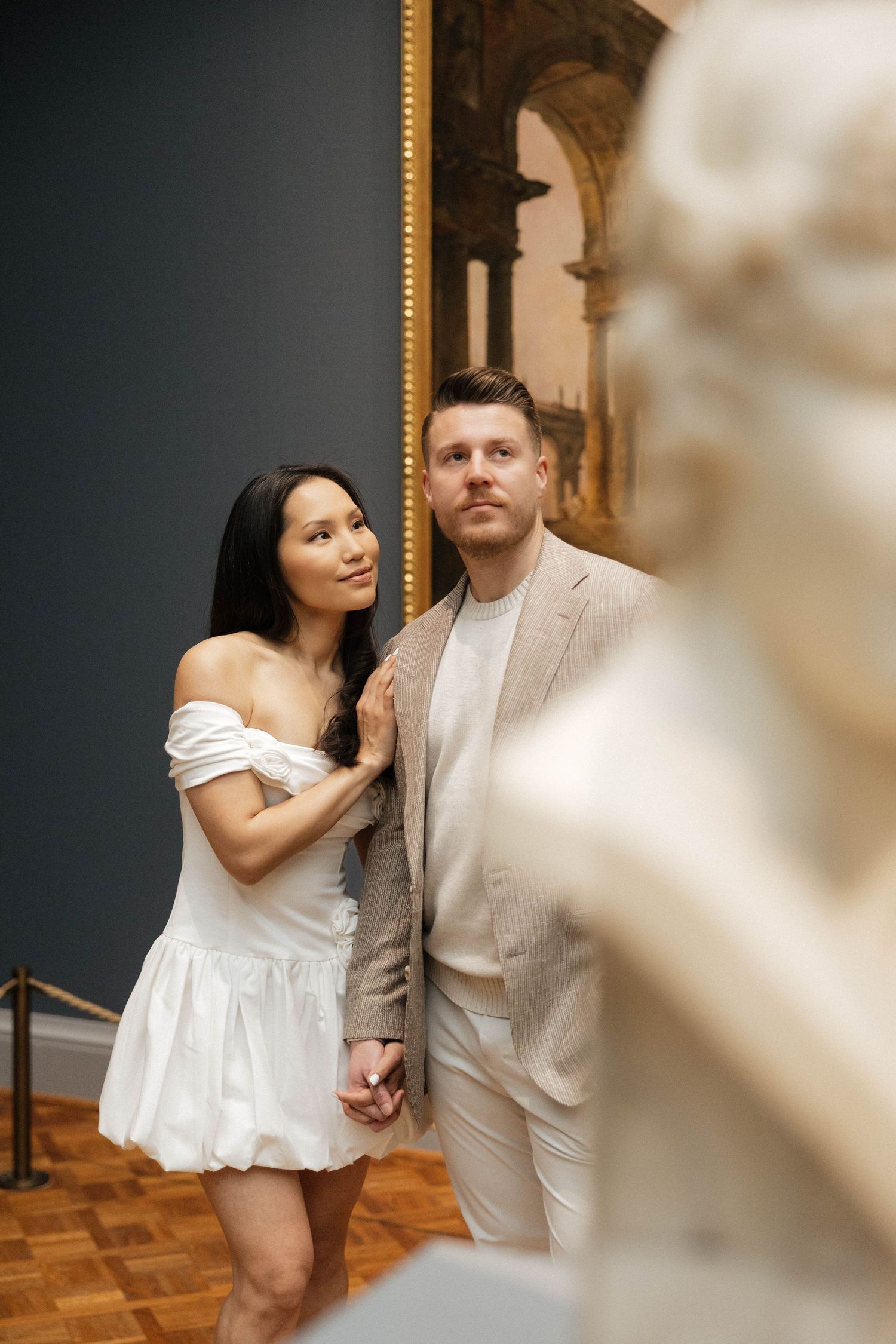 Couple holding hands in a museum, woman in white off-shoulder dress, man in beige suit, classic painting backdrop.
