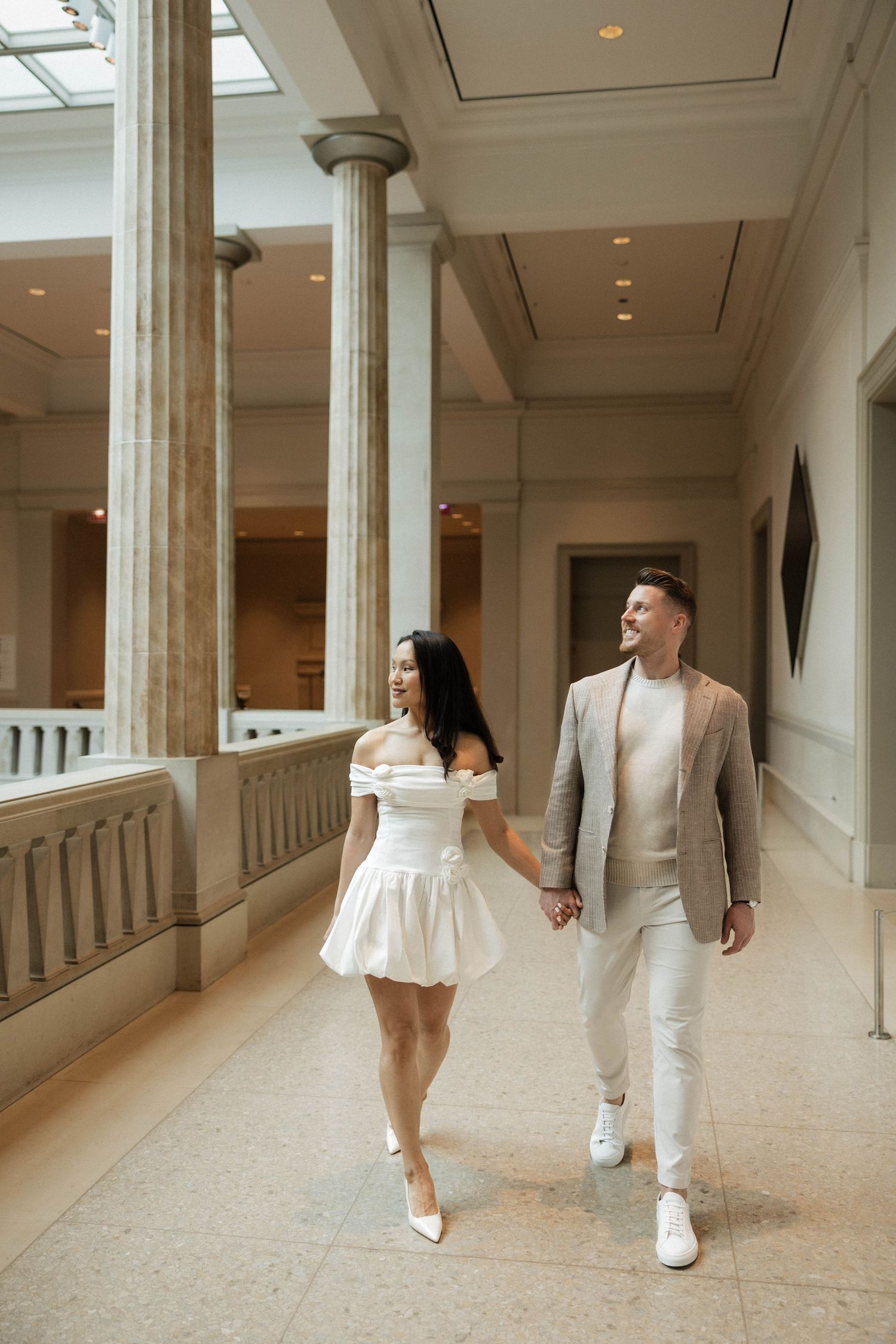 Elegant couple holding hands walking through a grand museum hallway with tall columns, wearing white and beige outfits.