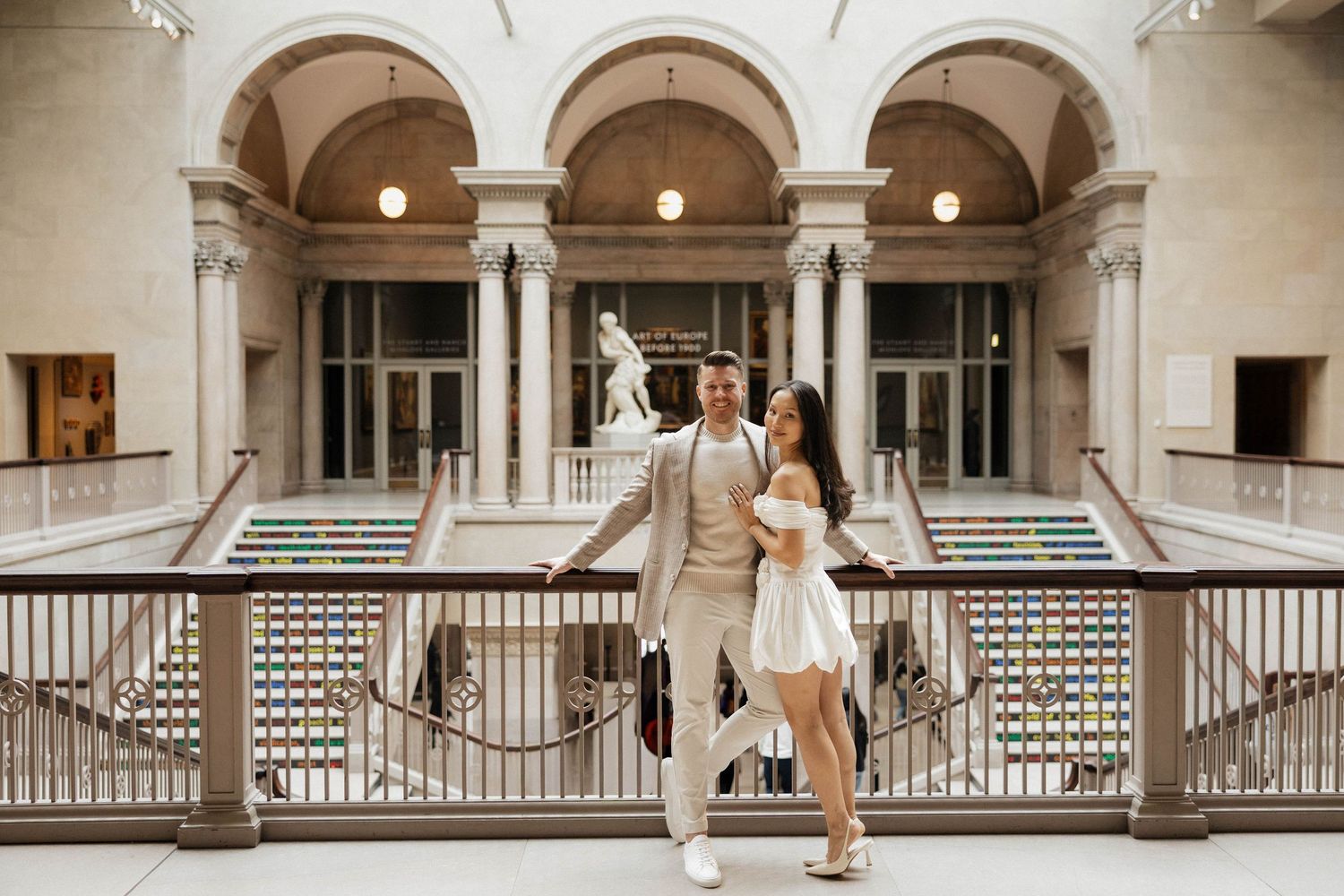 Couple posing elegantly on a balcony inside a grand museum with arched ceilings and marble staircases.