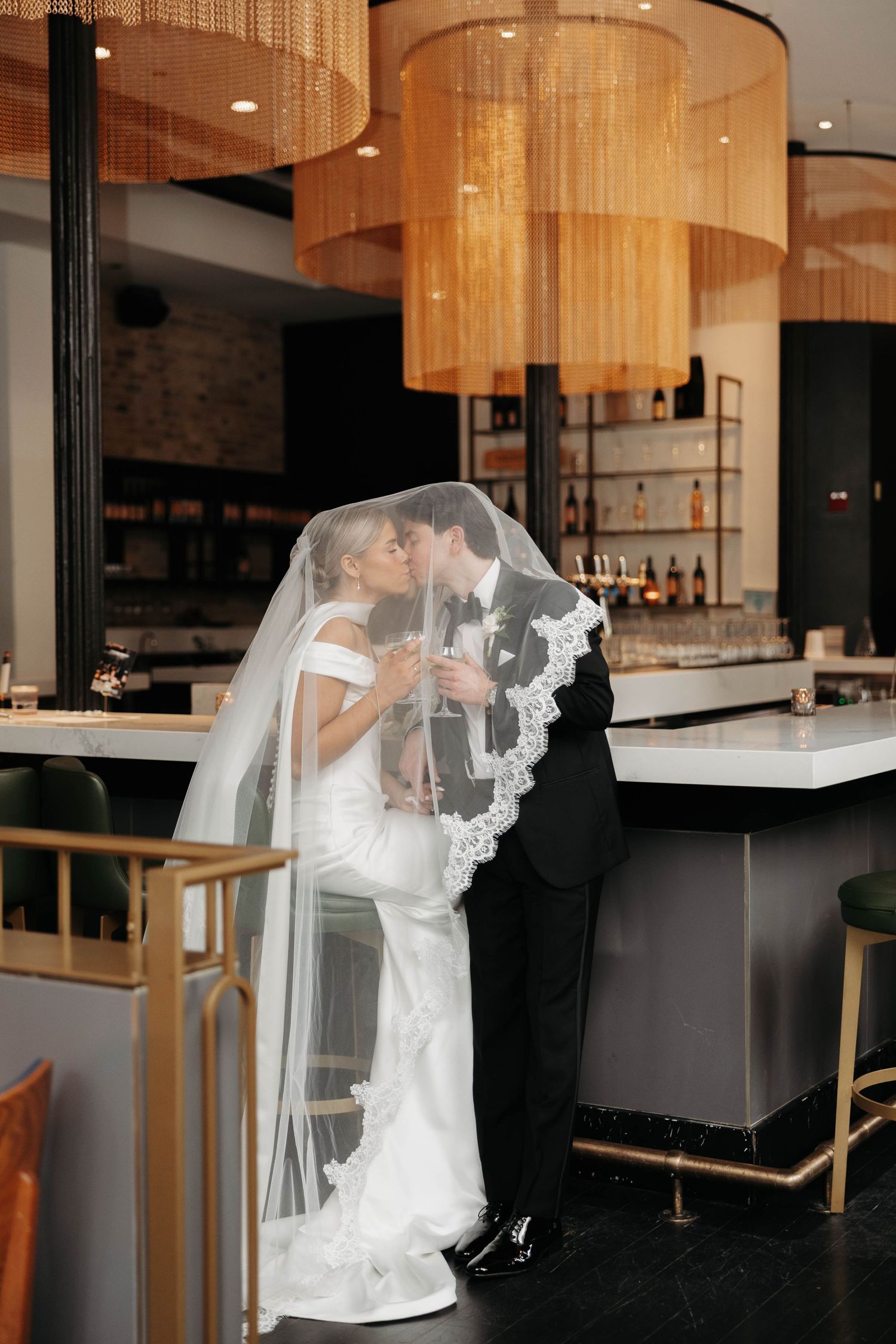Bride and groom kissing under veil at elegant bar with gold fringe chandeliers at Chicago Winery.