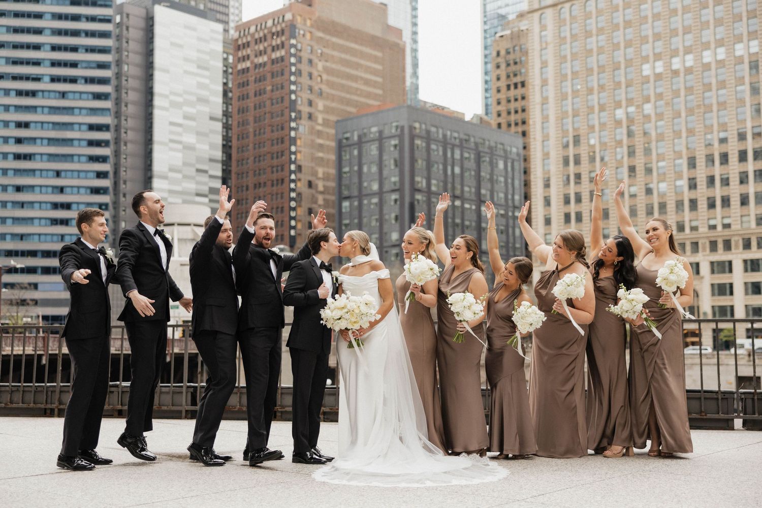 Wedding party celebrating on rooftop with city skyline, bridesmaids in gold gowns and groomsmen in black tuxedos.