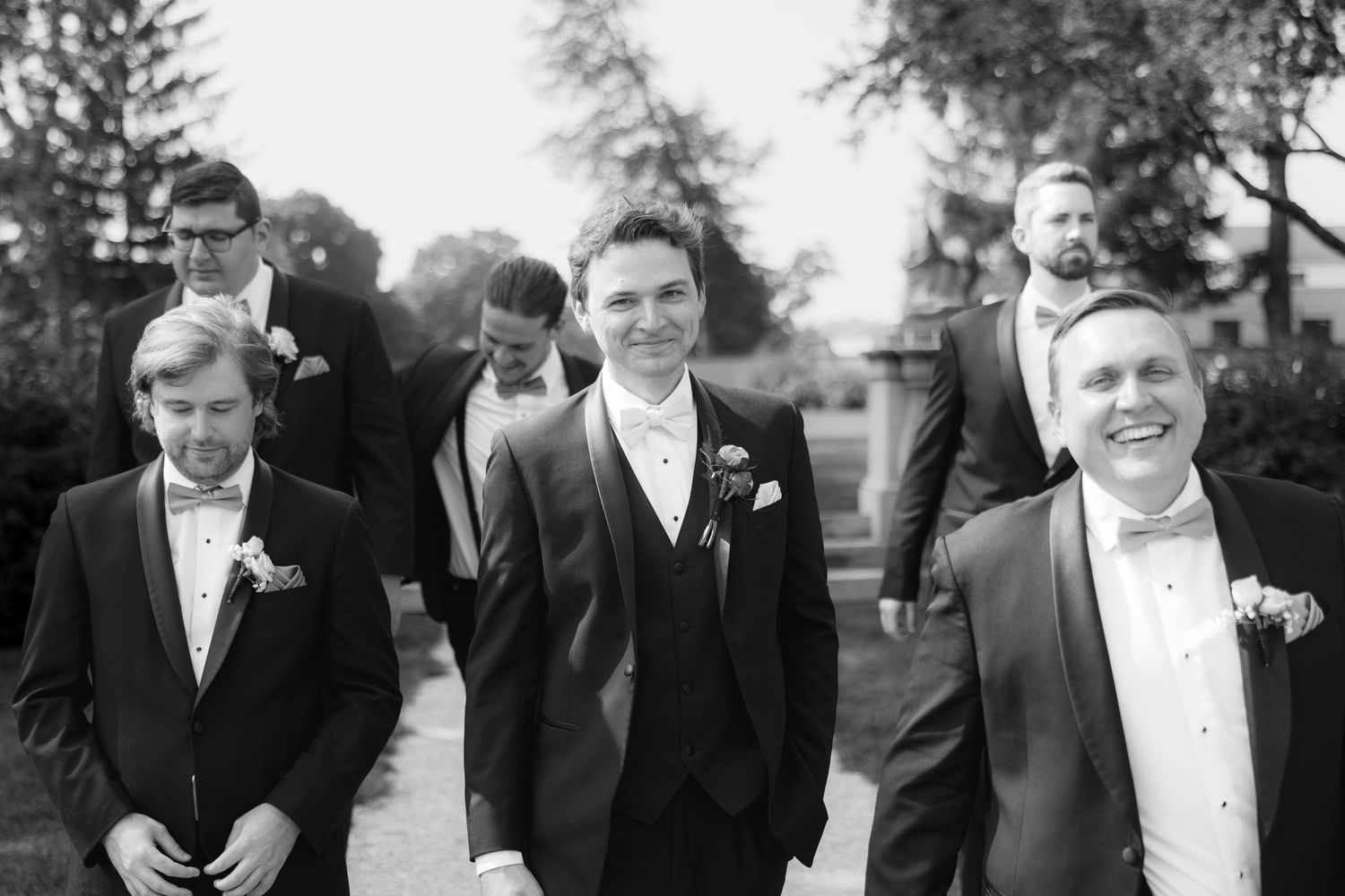 Groom and groomsmen in black tuxedos with bow ties walking together outdoors in a black and white wedding photo.