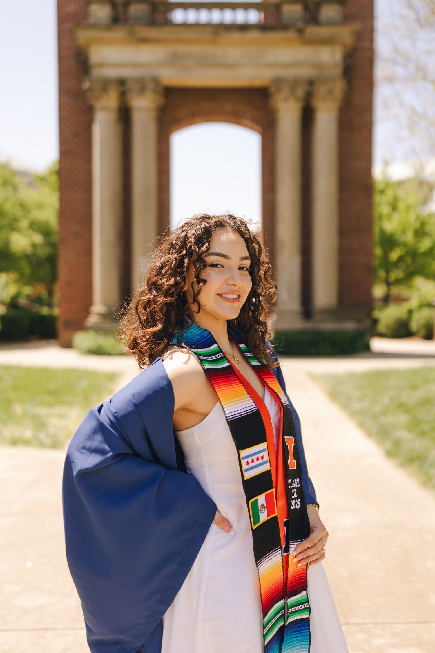 Graduate in blue cap and gown with colorful cultural stoles poses for graduation photos on a sunny day.
