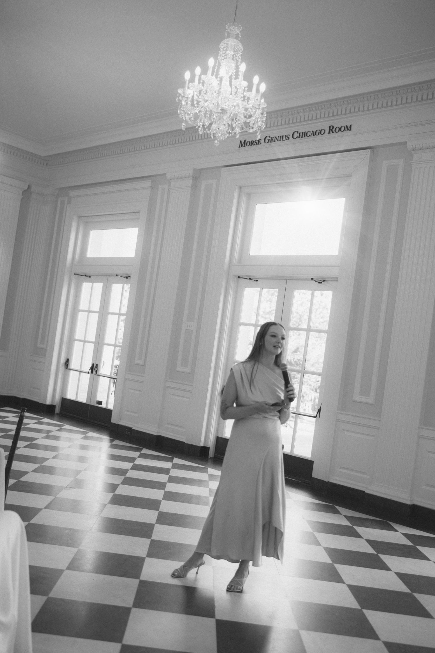 A figure stands in an elegant room with black and white checkered floors and a crystal chandelier overhead.