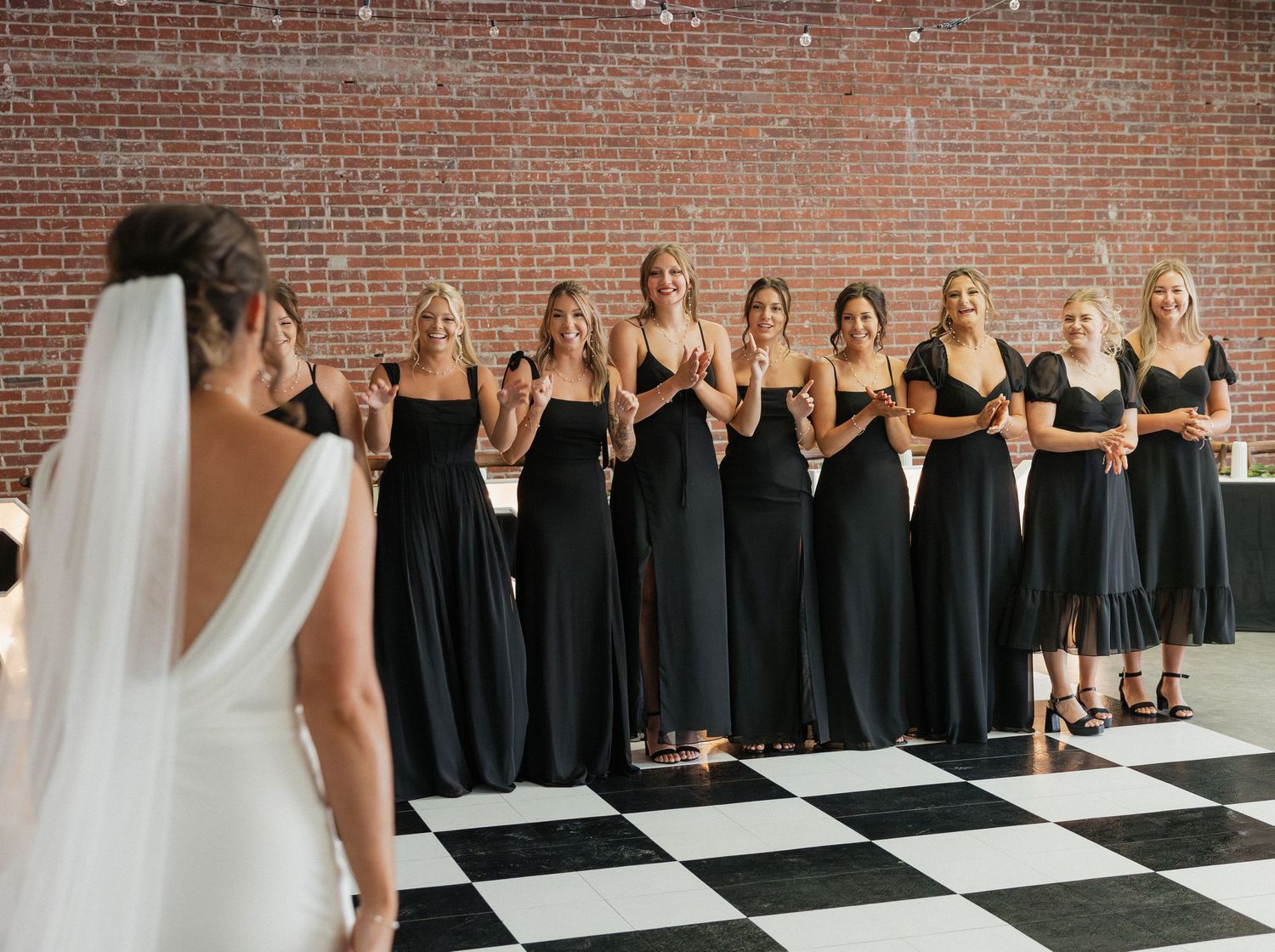 Wedding party poses on black and white checkered floor with bridesmaids in black dresses and bride in white gown.