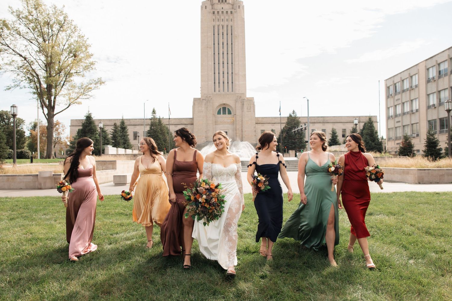 Wedding party members in colorful dresses pose together on a grassy lawn near a historic building.