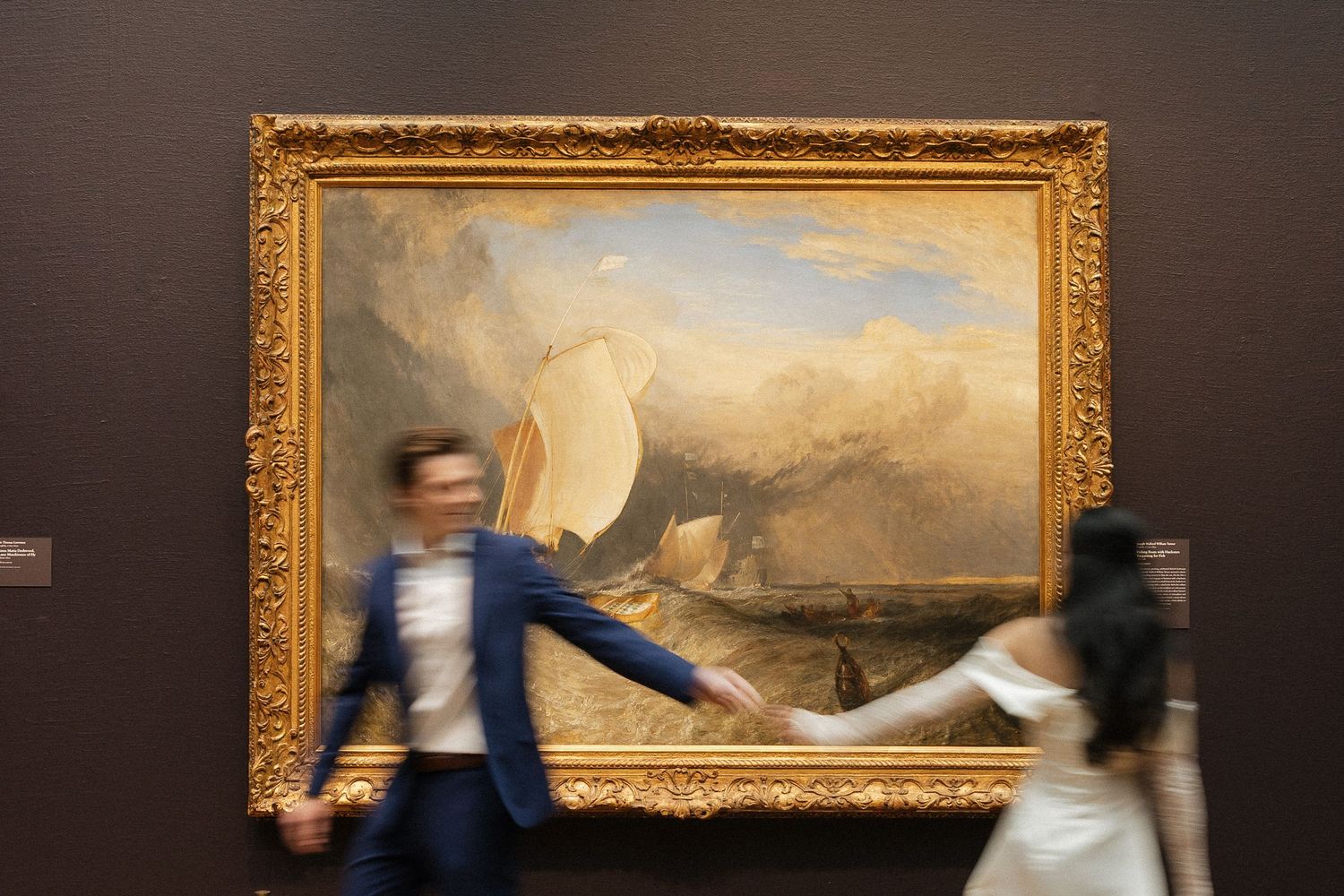 Art gallery interior with gold framed paintings and blurred figures moving past.