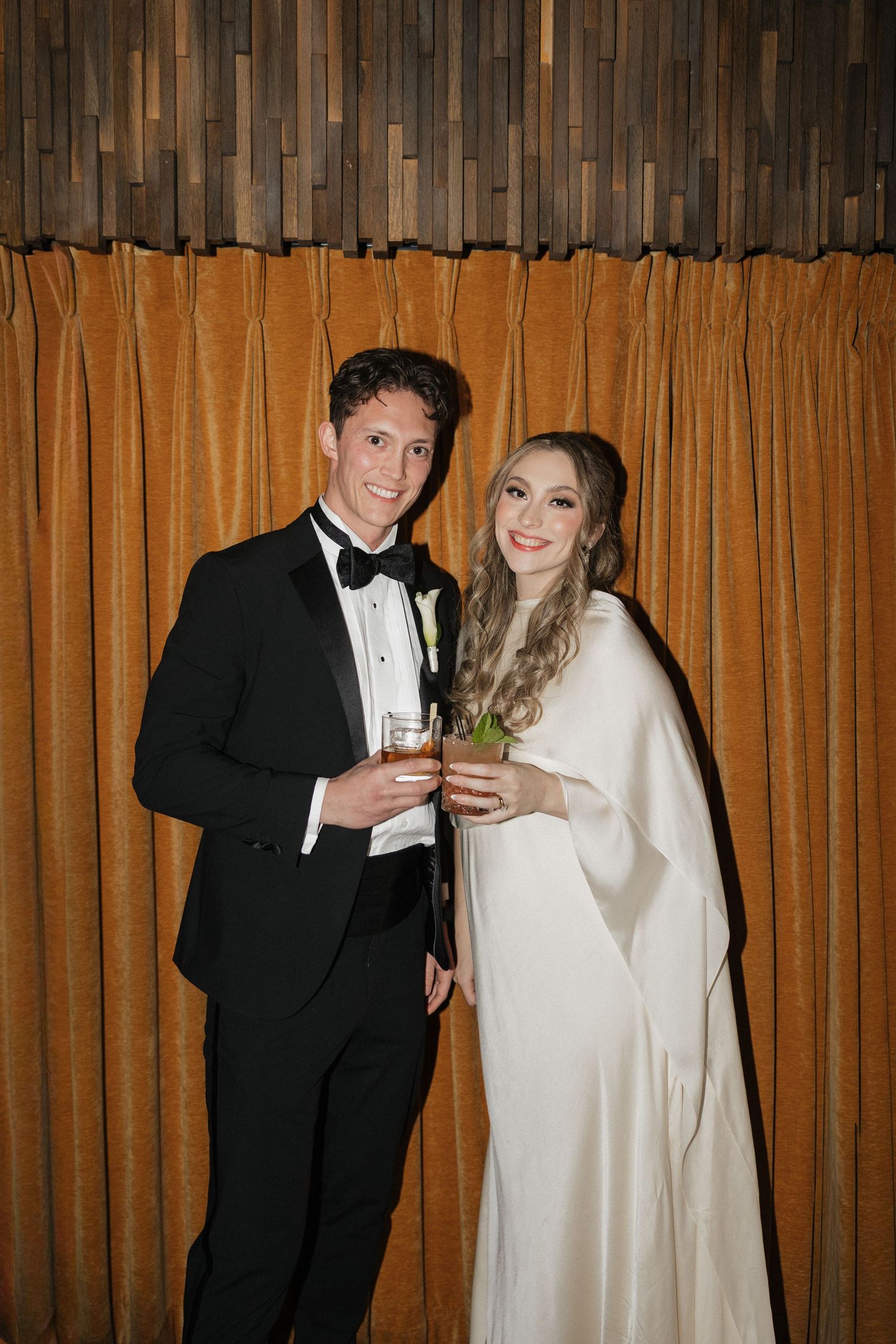 A couple in wedding attire sharing a drink together in front of orange curtains.