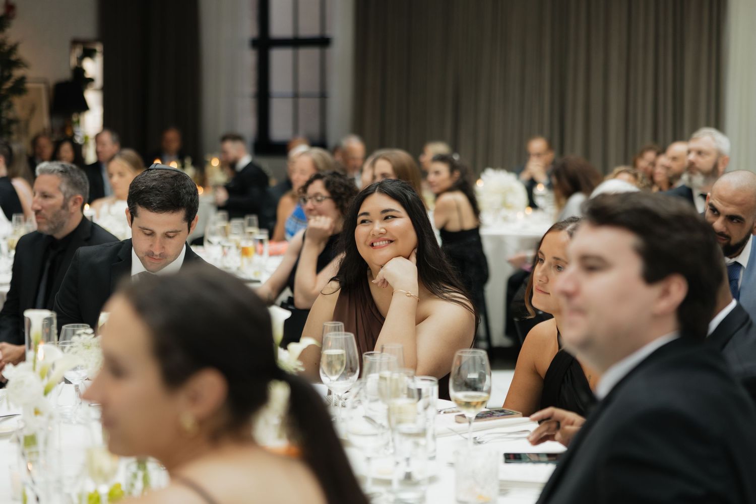 Wedding guests smile and laugh during reception speeches at elegantly set dinner tables.