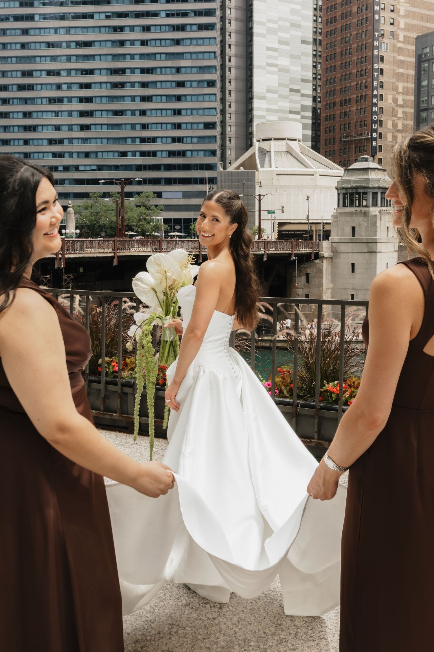 Bride in white gown holds bouquet with bridesmaids in brown dresses on urban rooftop with Chicago River backdrop.