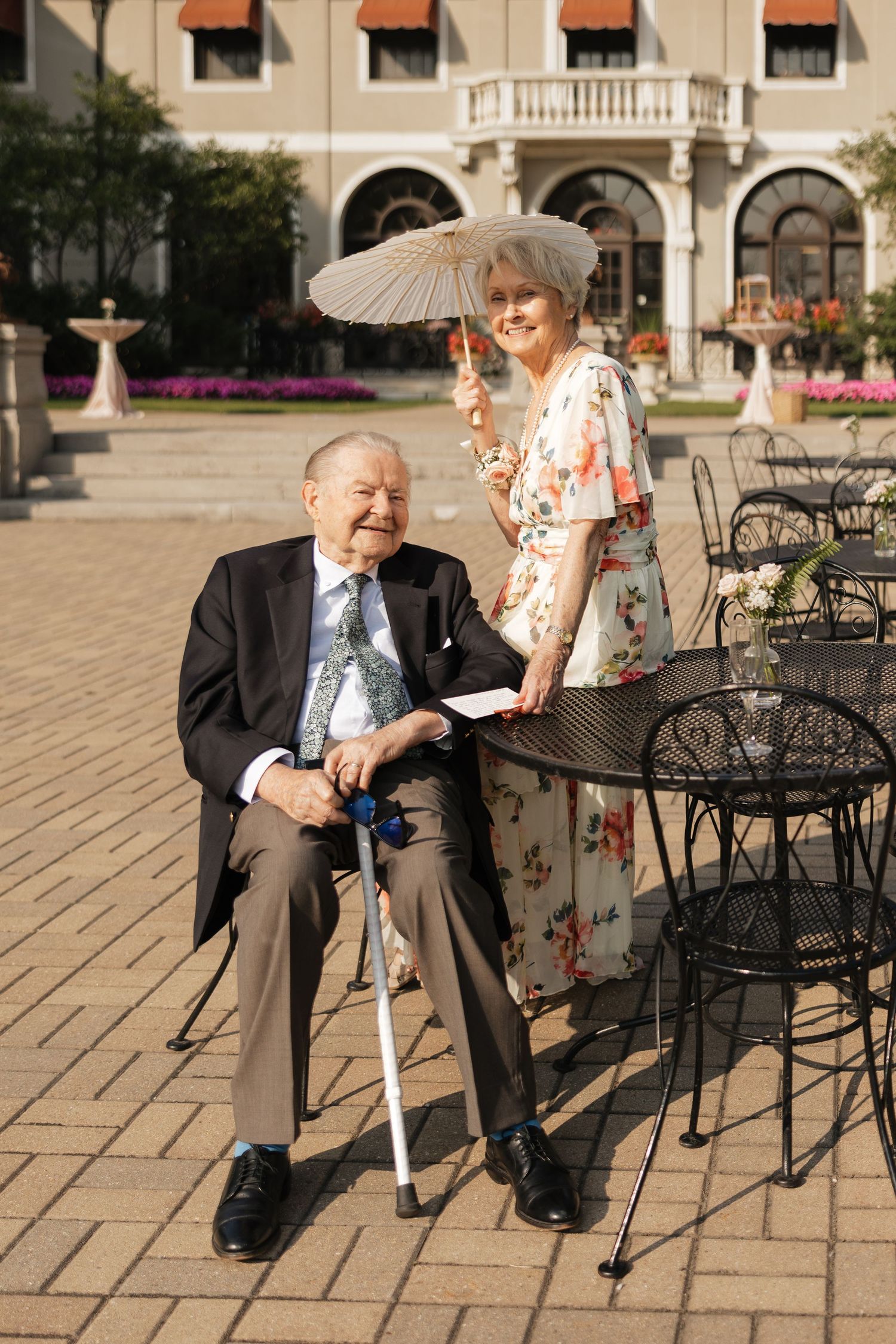 Elderly man with cane seated outdoors beside woman holding parasol near elegant building with iron chairs.