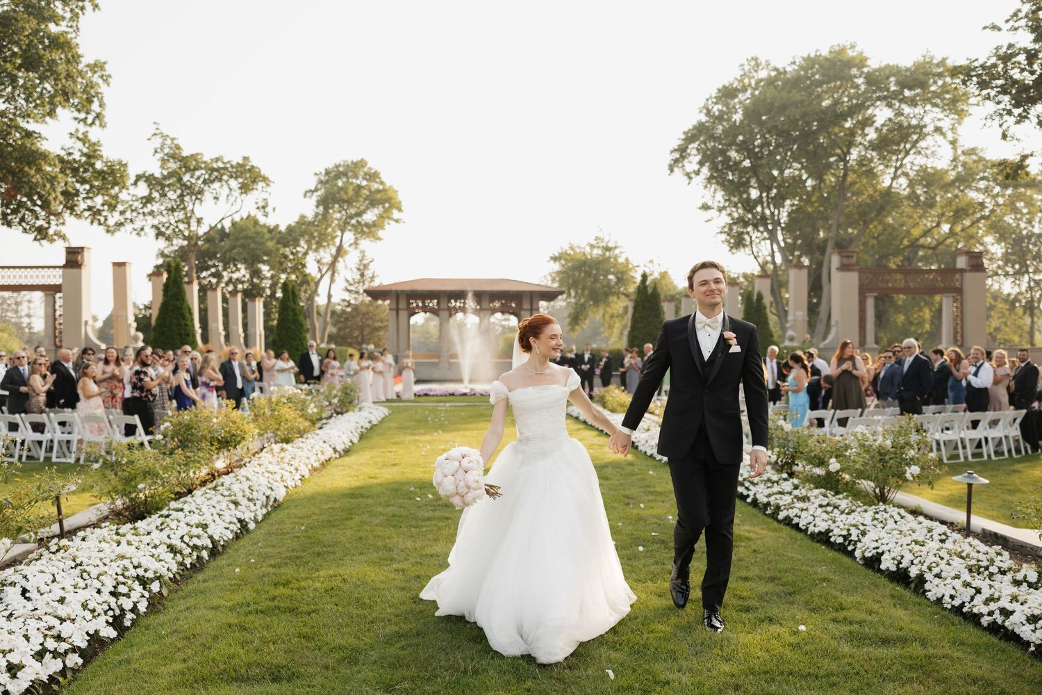 Bride and groom walk hand-in-hand down a floral-lined garden aisle with a fountain and guests in the background.