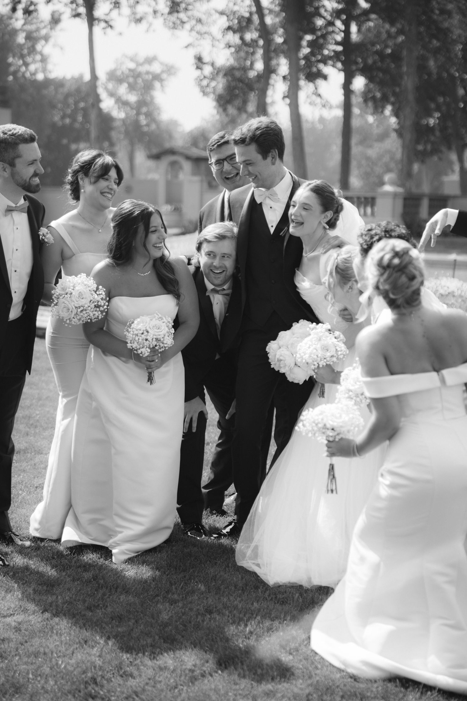 Black and white photo of joyful wedding party laughing together on lawn, bridesmaids holding white floral bouquets.