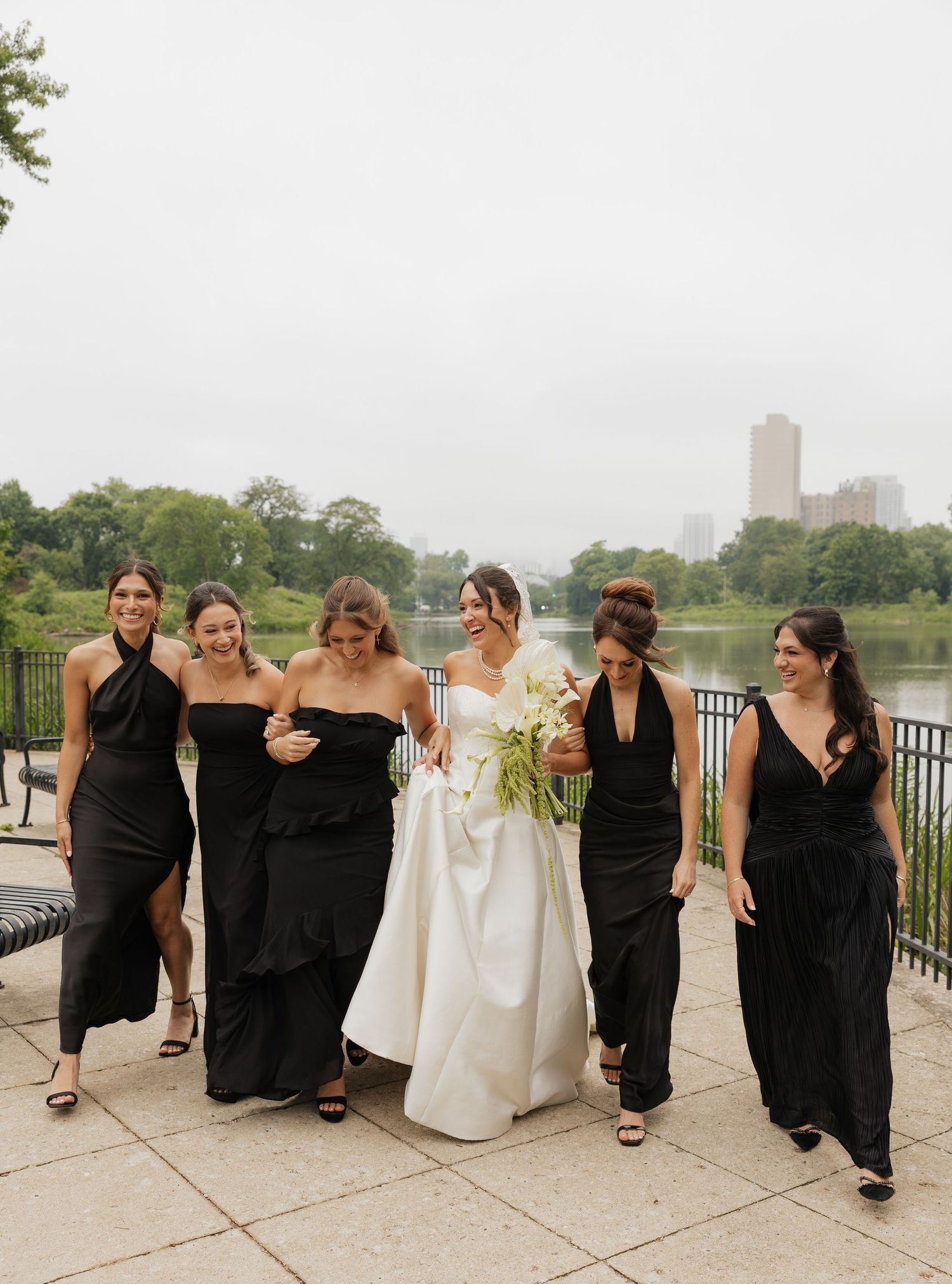 Bride in white gown walks with five bridesmaids in black dresses near a park lake on a cloudy day.