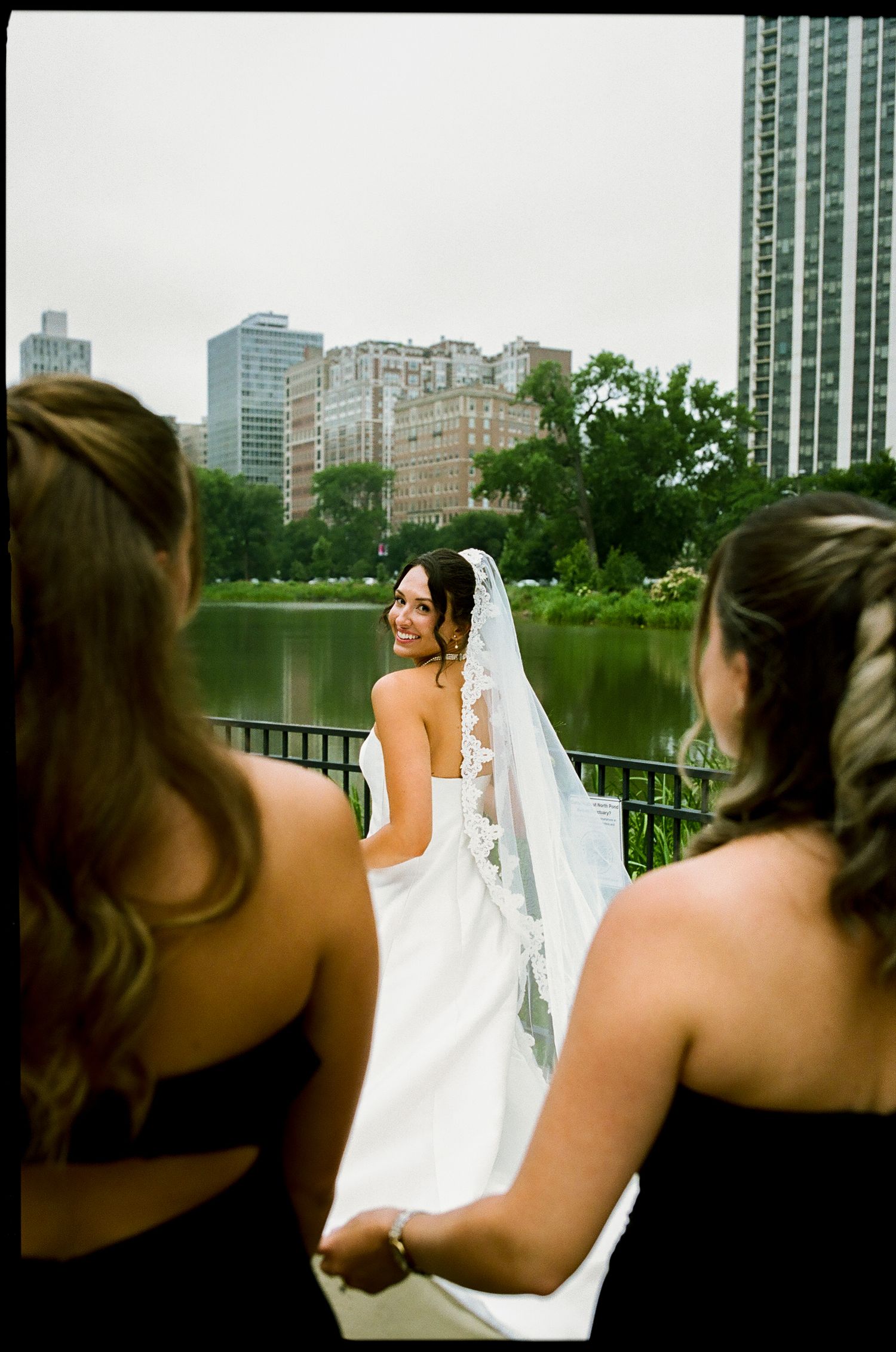 Smiling bride in white lace veil gown stands on bridge with bridesmaids in black dresses, city skyline behind.