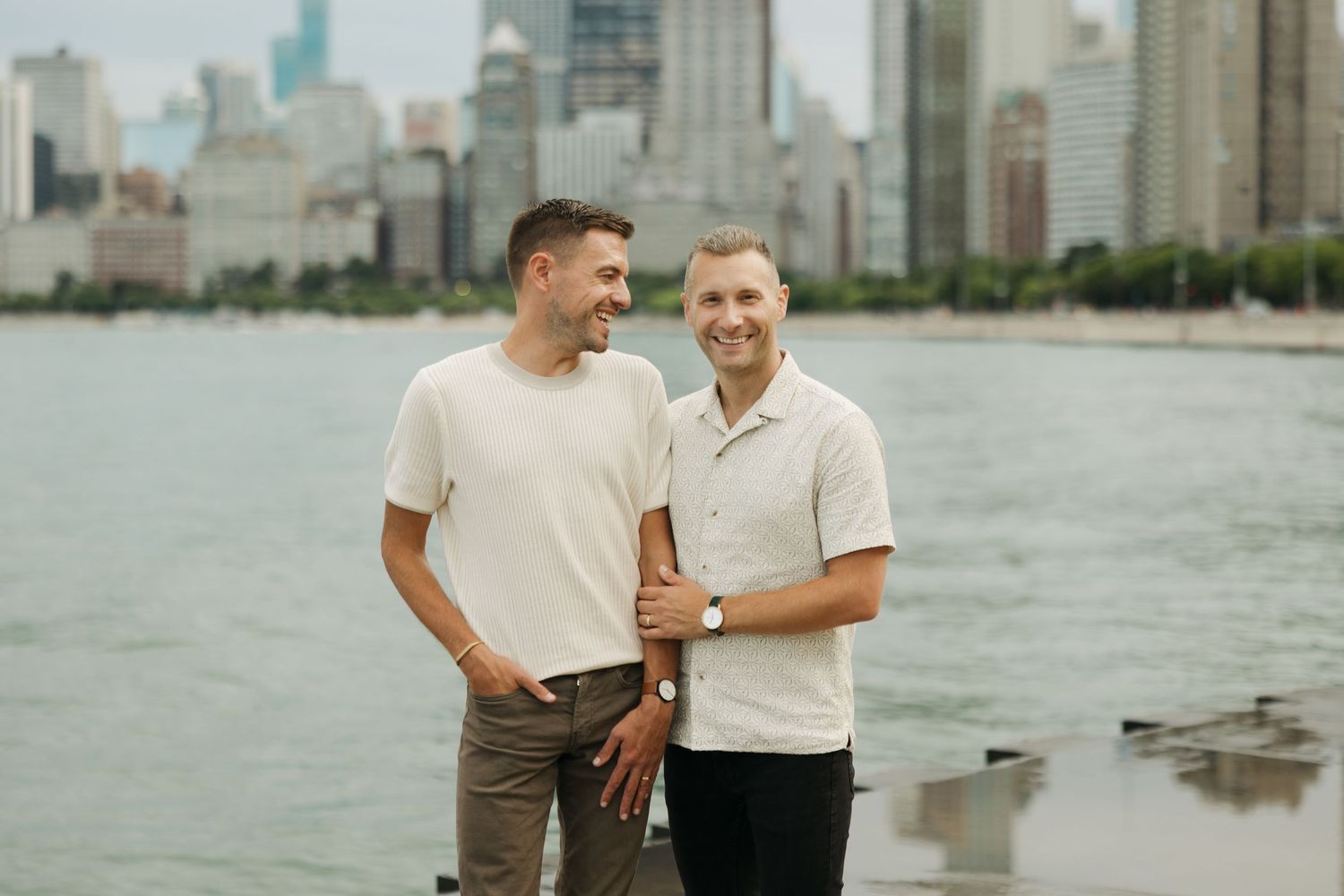 Two smiling men in light casual shirts stand close together by a lakefront with a hazy city skyline rising behind them.