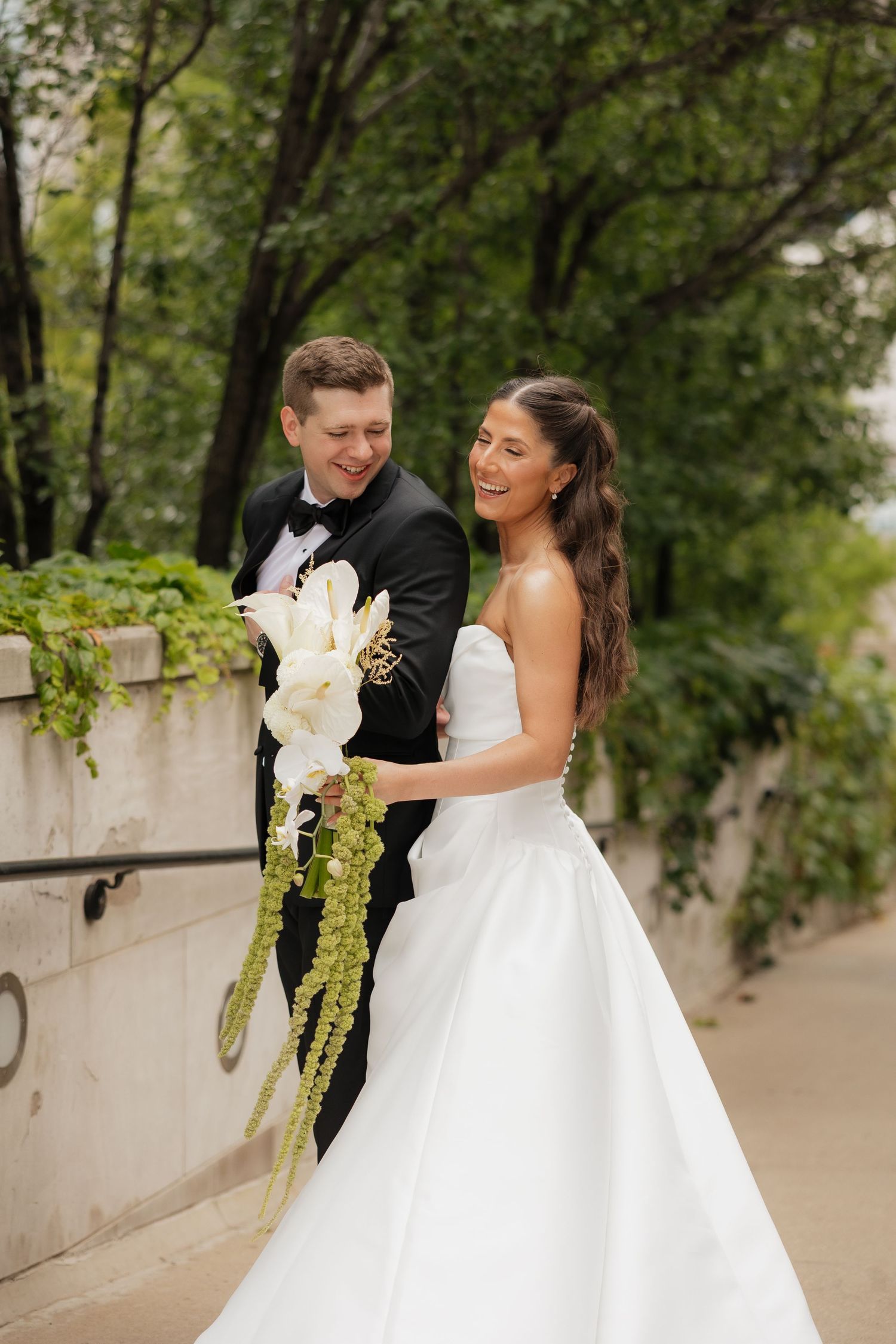 Smiling bride in a strapless white gown holding a white orchid and green amaranthus bouquet beside her laughing groom in a black tuxedo.