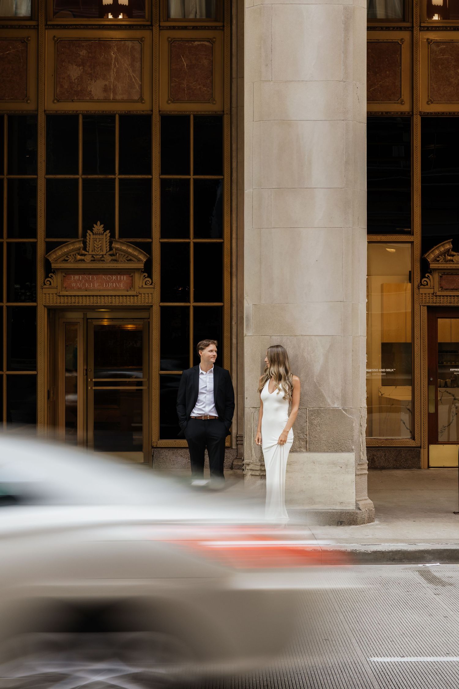 Couple in formal attire stands beside a stone column outside an ornate building entrance while a blurred car speeds by on the city street.